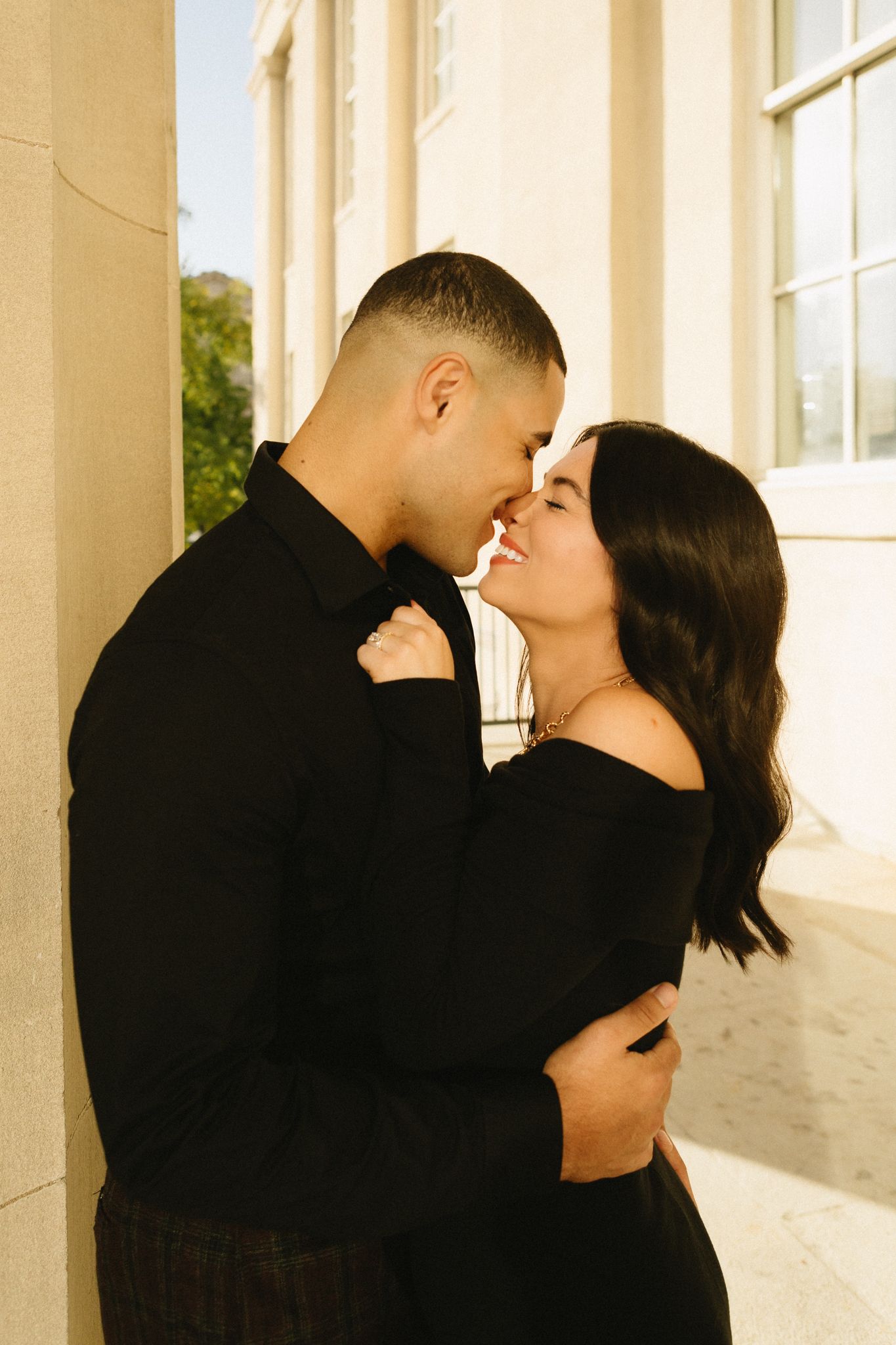 A tender moment between a couple in black attire against a light-colored building exterior.
