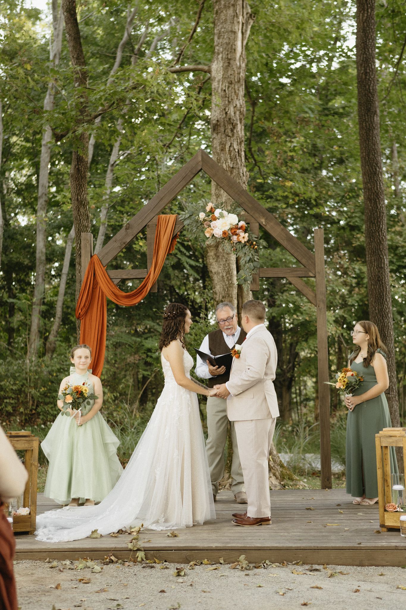Wedding ceremony taking place on a rustic wooden deck surrounded by trees with orange fabric draping decorations.