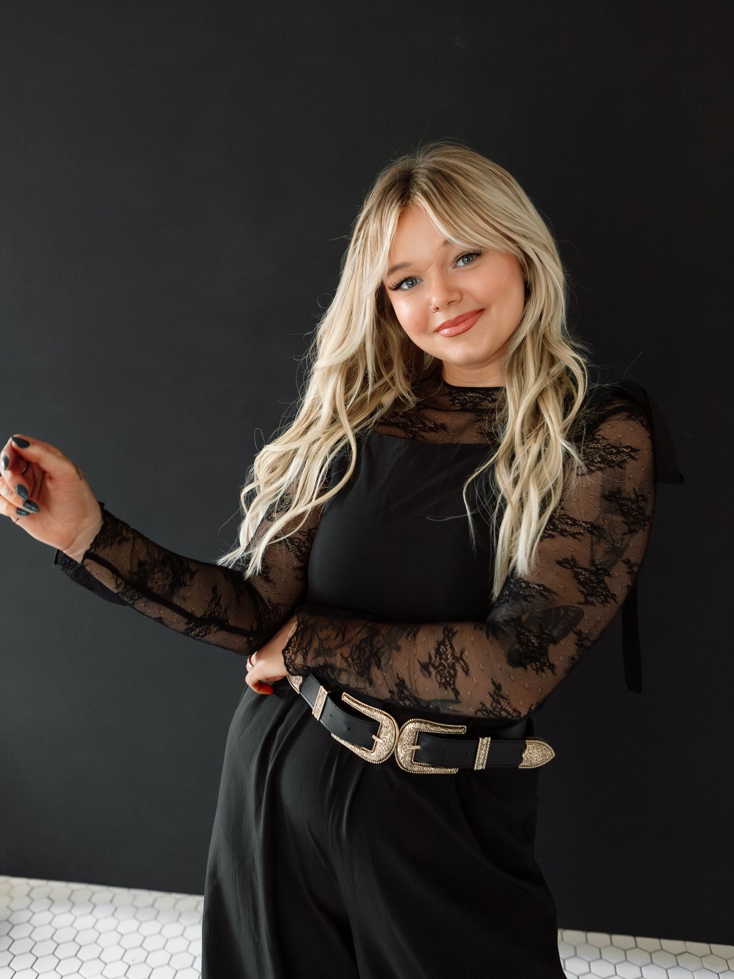 A smiling blonde woman in a black lace top and belt poses against a dark gray background.