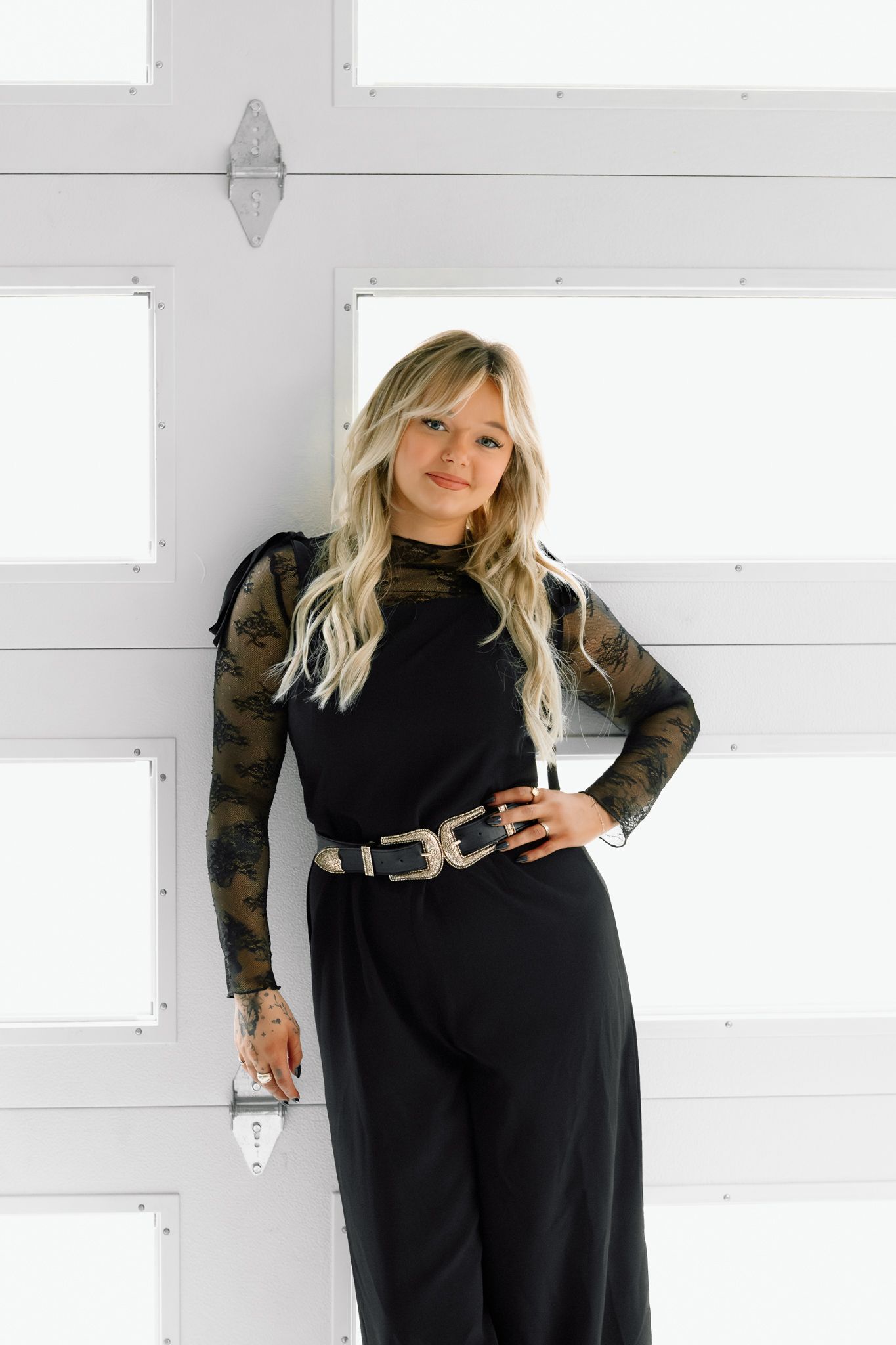 Blonde woman posing in black jumpsuit with camo mesh sleeves and gold belt against white garage door.