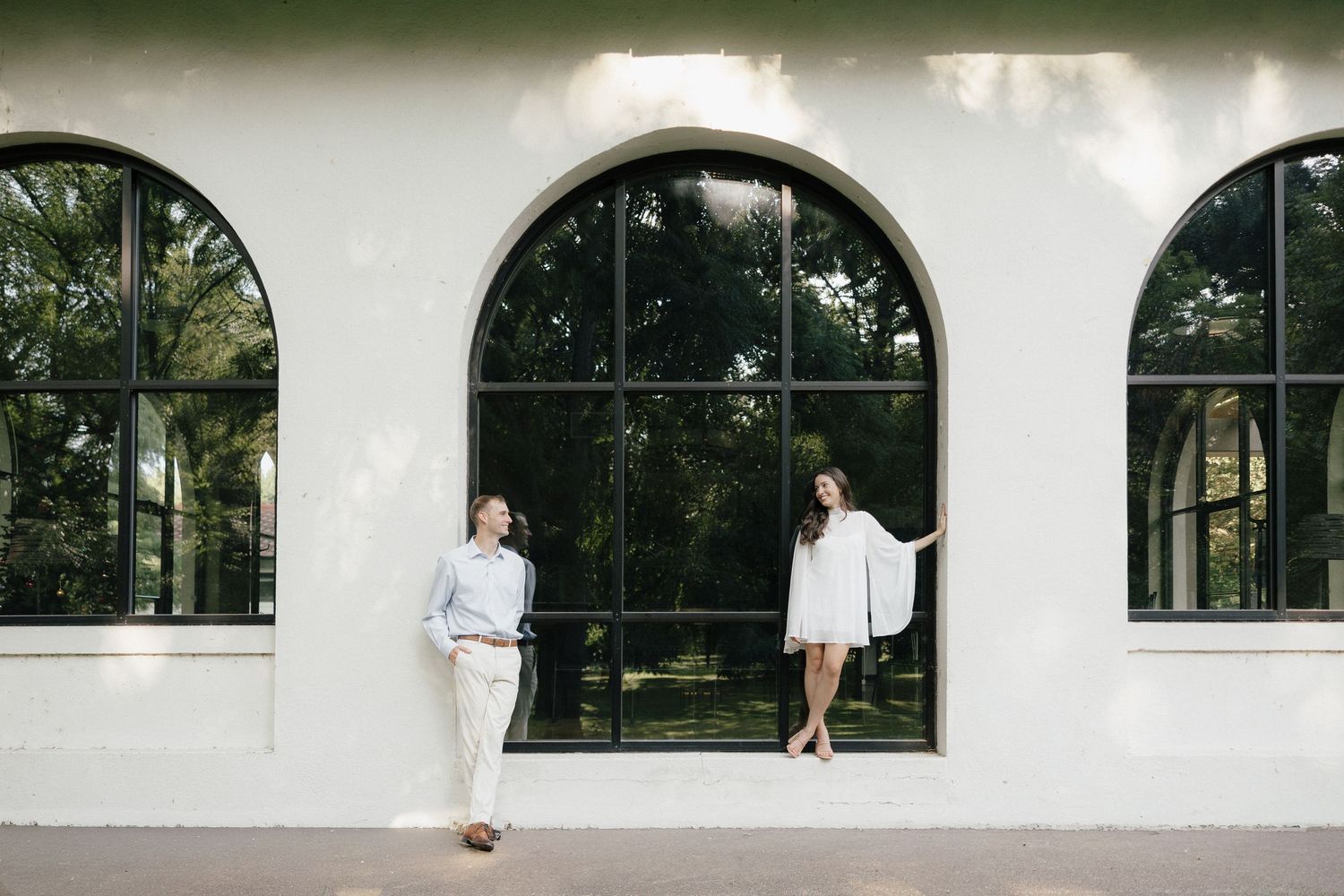 Engaged couple posing apart in front of a white building with large arched black-framed windows.