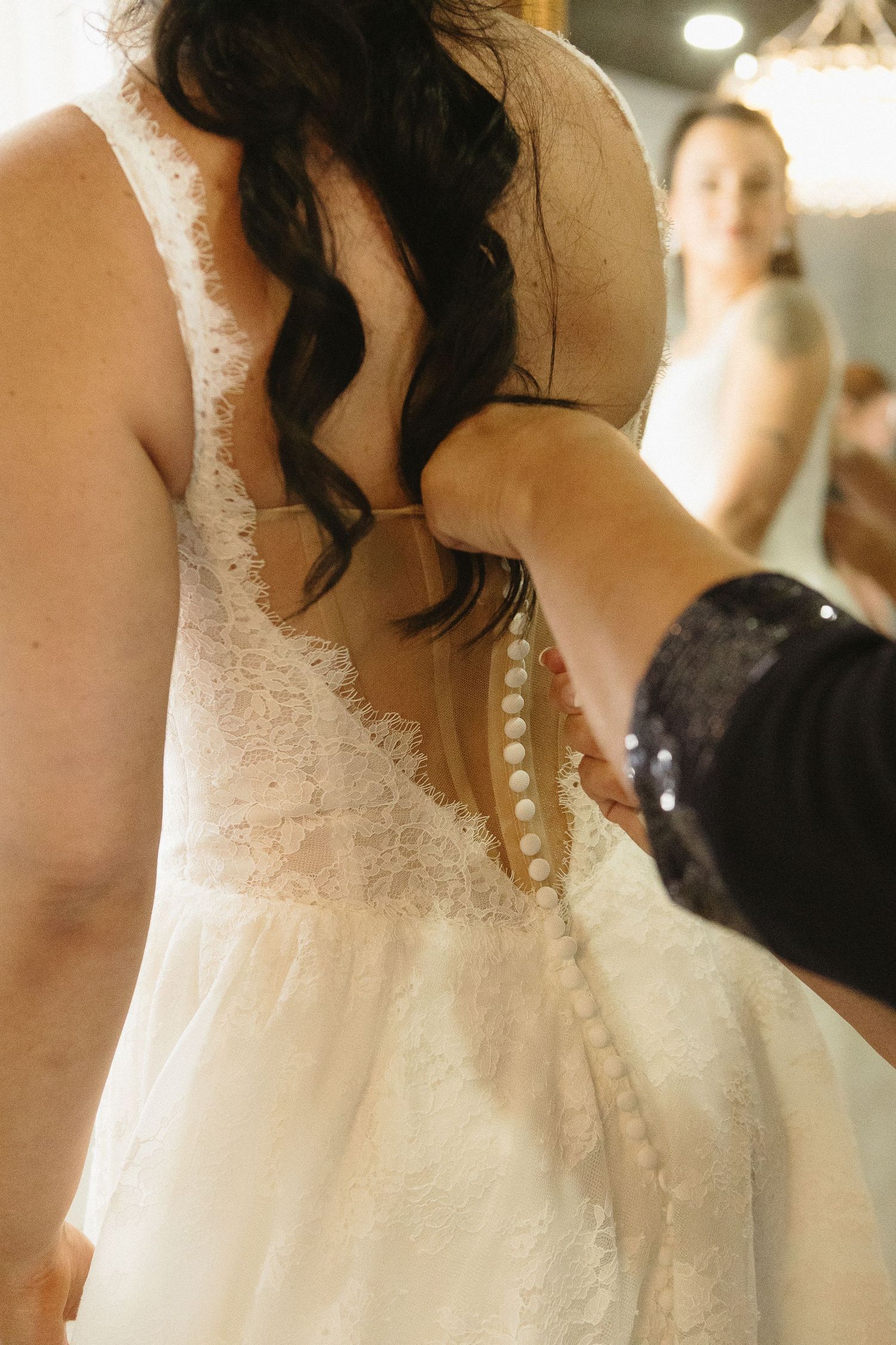 Someone helps button up a delicate white lace wedding dress with pearl buttons down the back.
