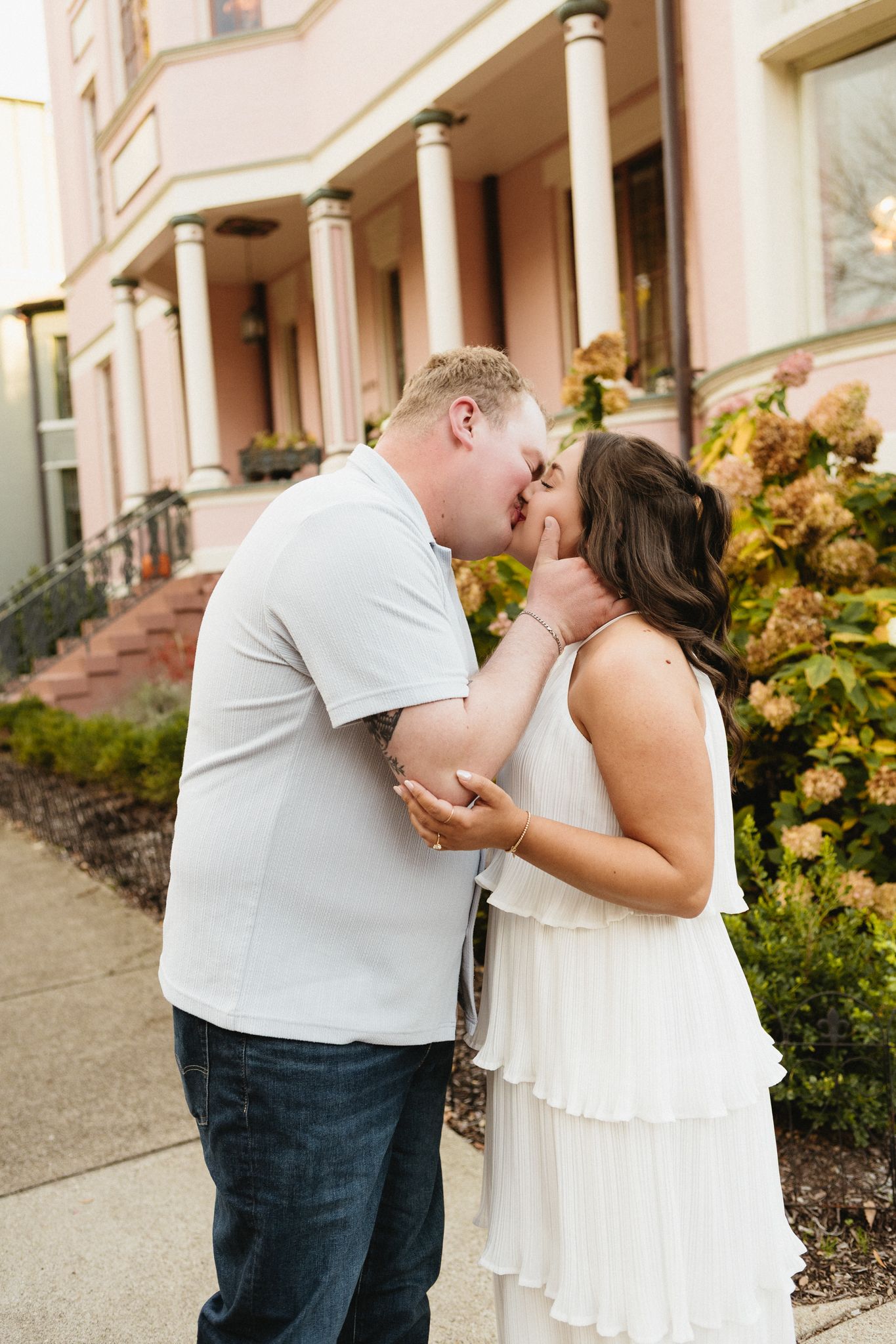 A romantic kiss shared on the steps of a pink columned building with hydrangeas blooming nearby.