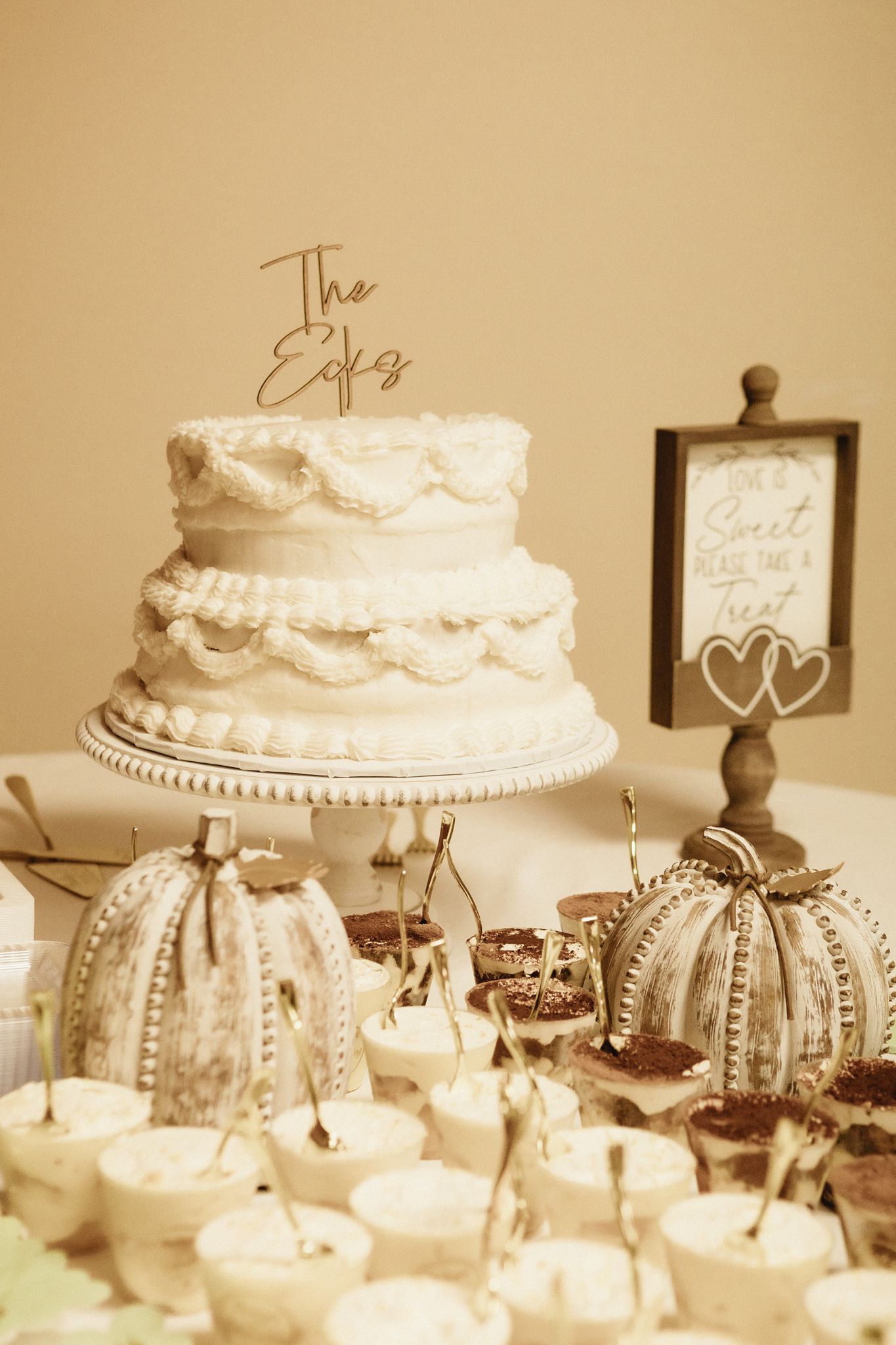 Elegant white wedding cake and cupcake display with vintage-style decorations and framed signage.