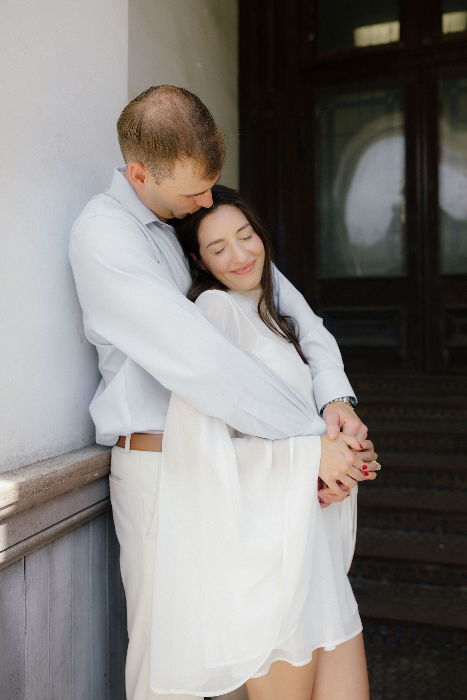 Man tenderly embracing woman in white dress against elegant architectural column in soft light.