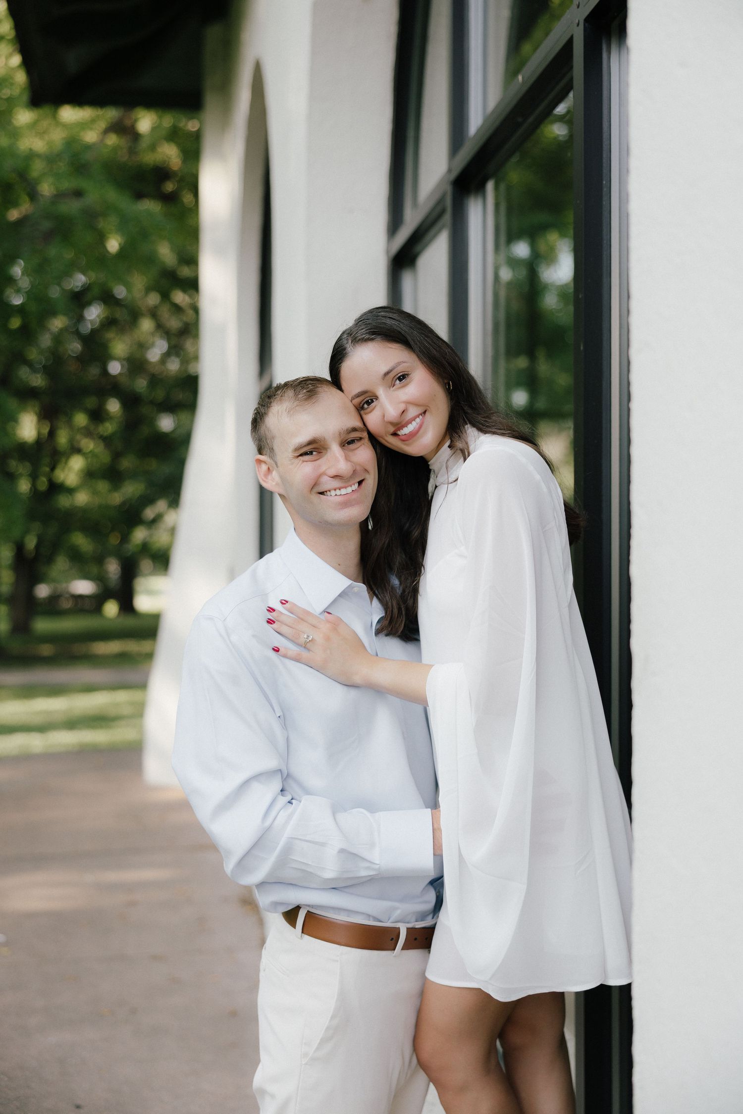 A smiling couple in white outfits embracing outdoors near a building, woman resting her head on mans shoulder.