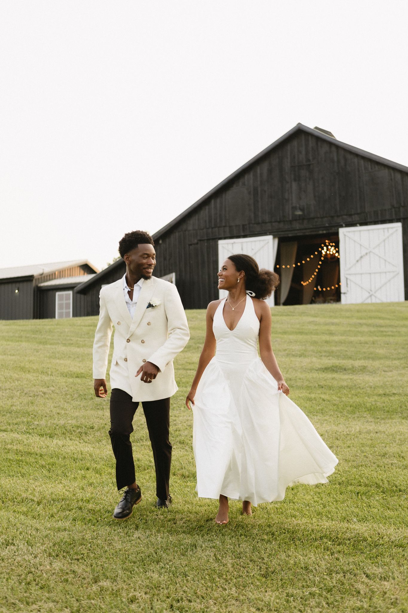 A couple dances playfully in front of a rustic barn on a grassy field.
