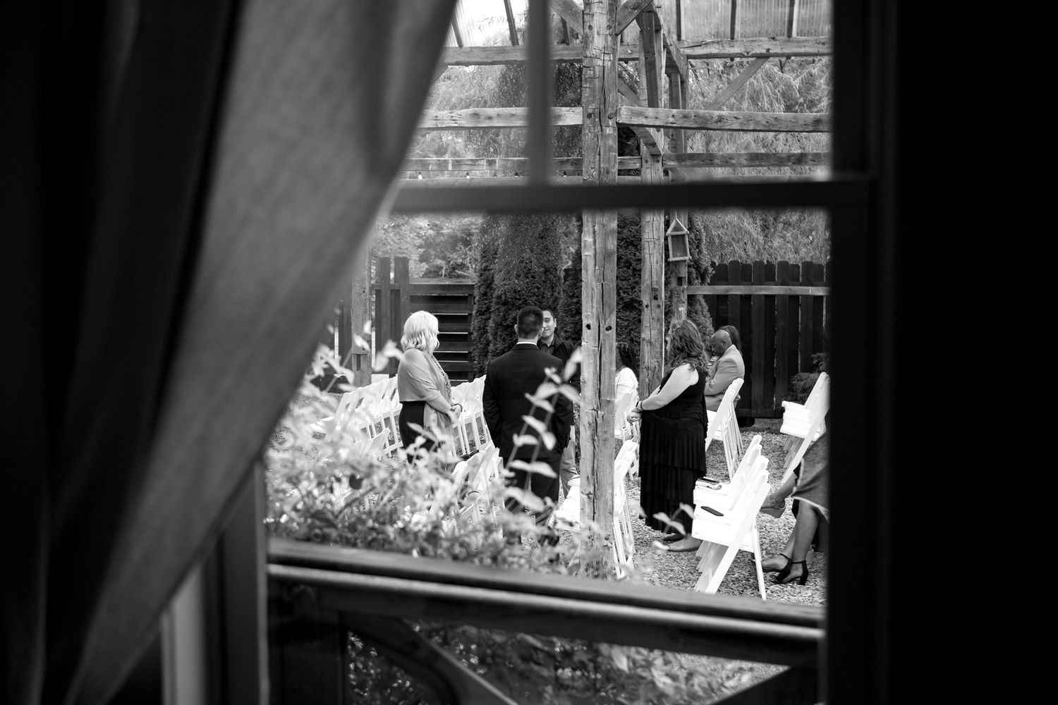 A black and white view through a window of outdoor party guests gathered in a garden setting.