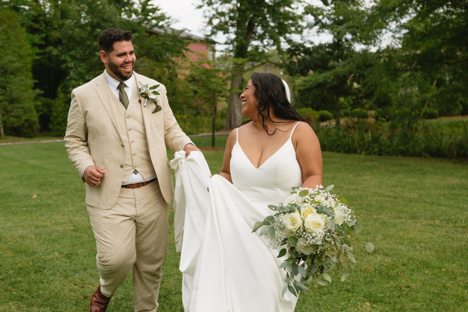 Couple in wedding attire walking together on green lawn with bride carrying white and green floral bouquet.