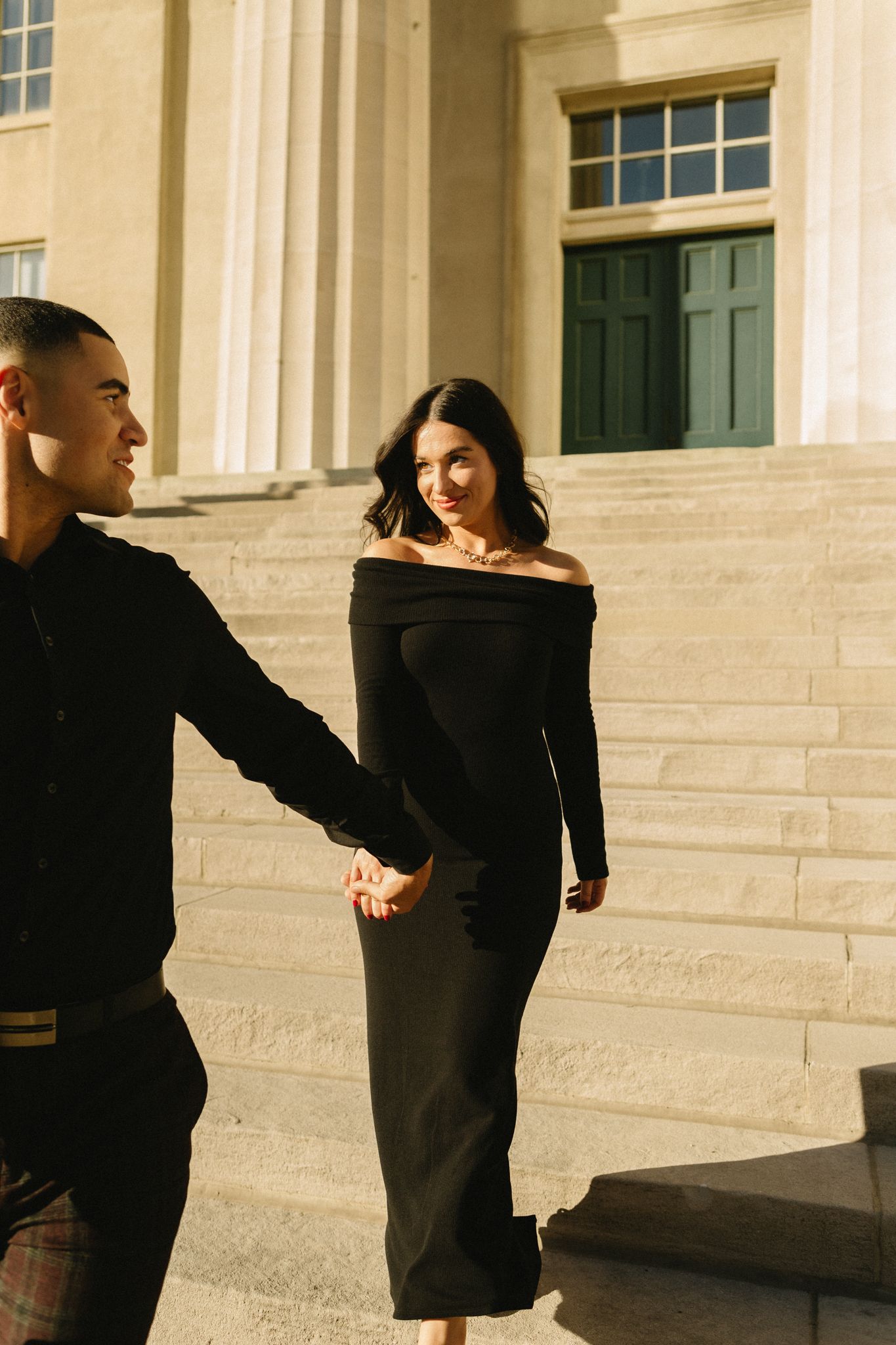 A couple in matching black outfits dance together on stone steps against a classical building facade.