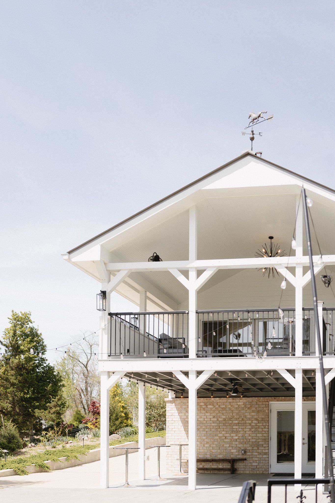 Modern two-story white pavilion with weather vane features exposed beams and covered balcony overlooking scenic outdoor area.