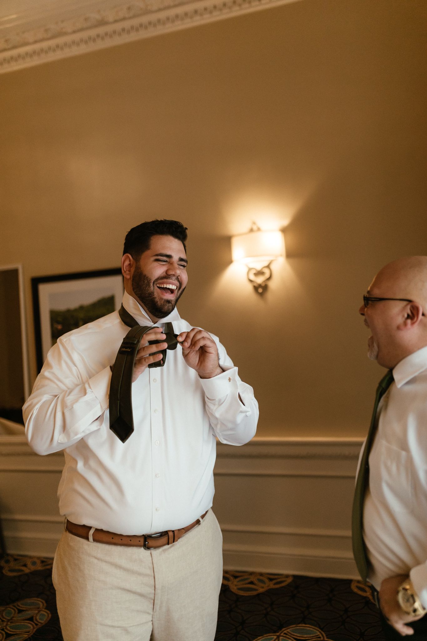 Two people dressed in formal white shirts and ties share a candid moment laughing together in an elegant room.