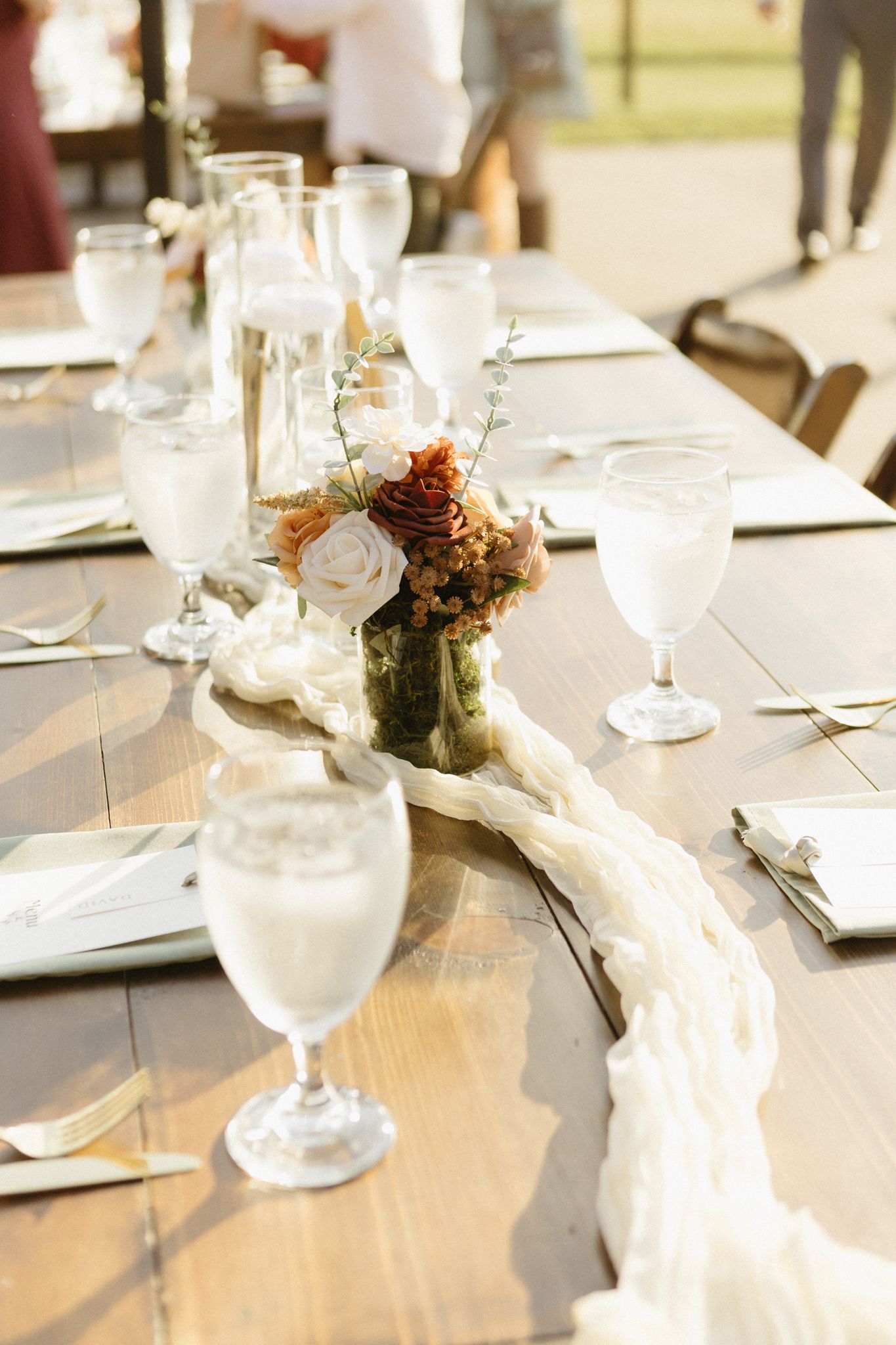 Elegant wedding table setting with white linens, wine glasses and small floral centerpieces on a wooden table.