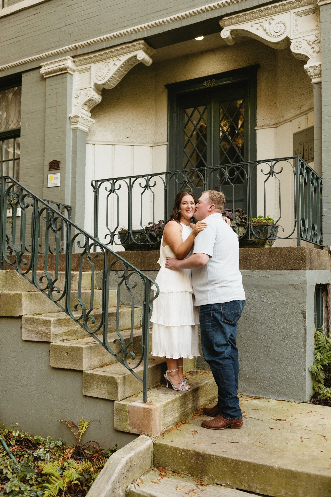 Two people share an intimate moment on the front steps of a classic brownstone with decorative metalwork.