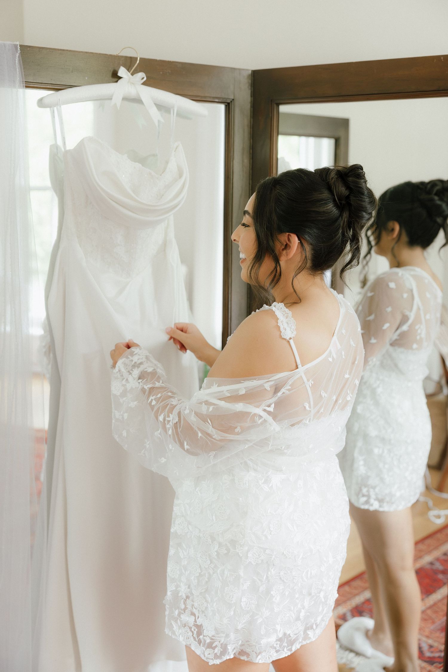 Bride in lace robe admiring her white wedding gown hanging by a sunlit window before her ceremony.