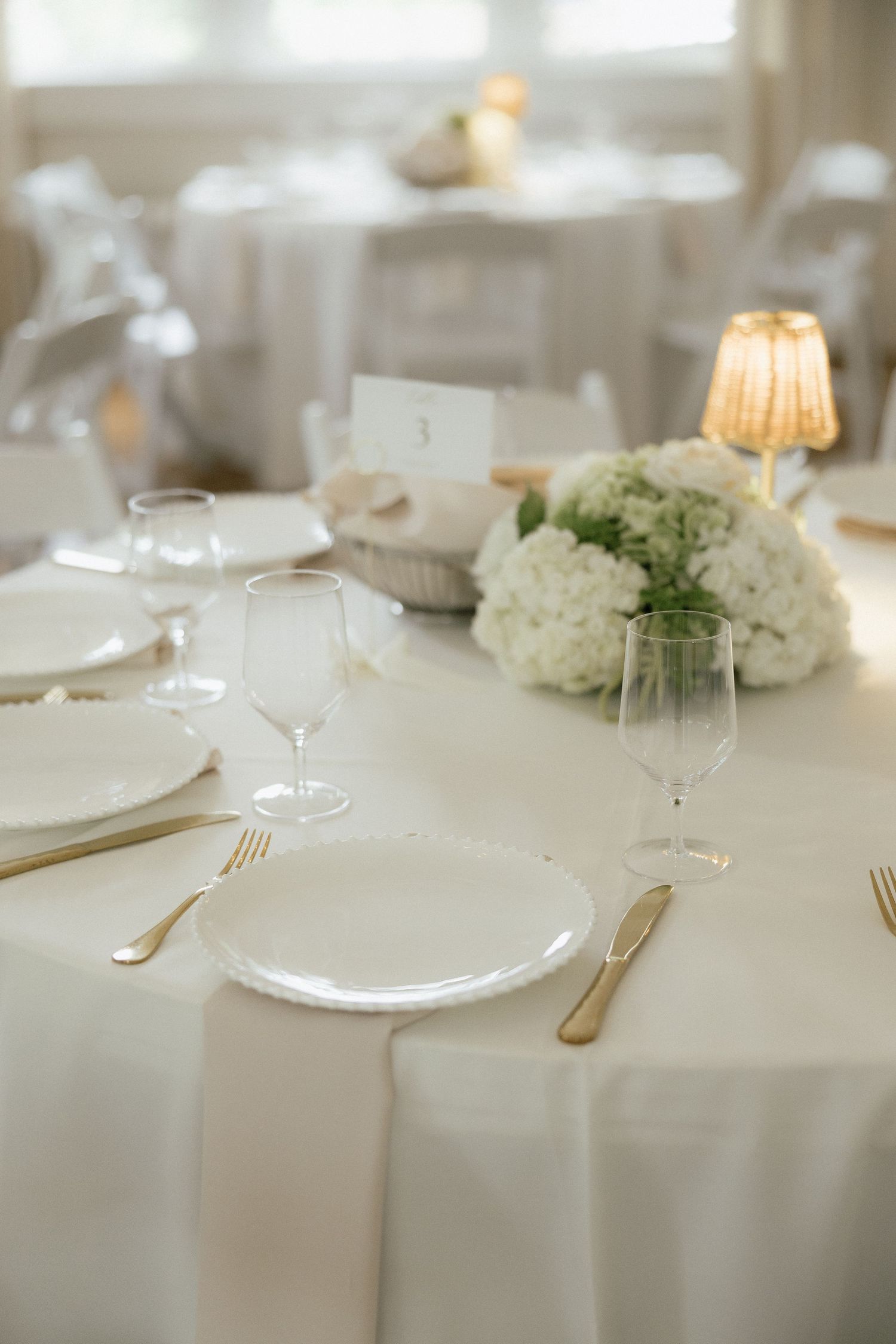 Elegant white wedding reception table setting with gold flatware, crystal glasses, and white hydrangea centerpiece.