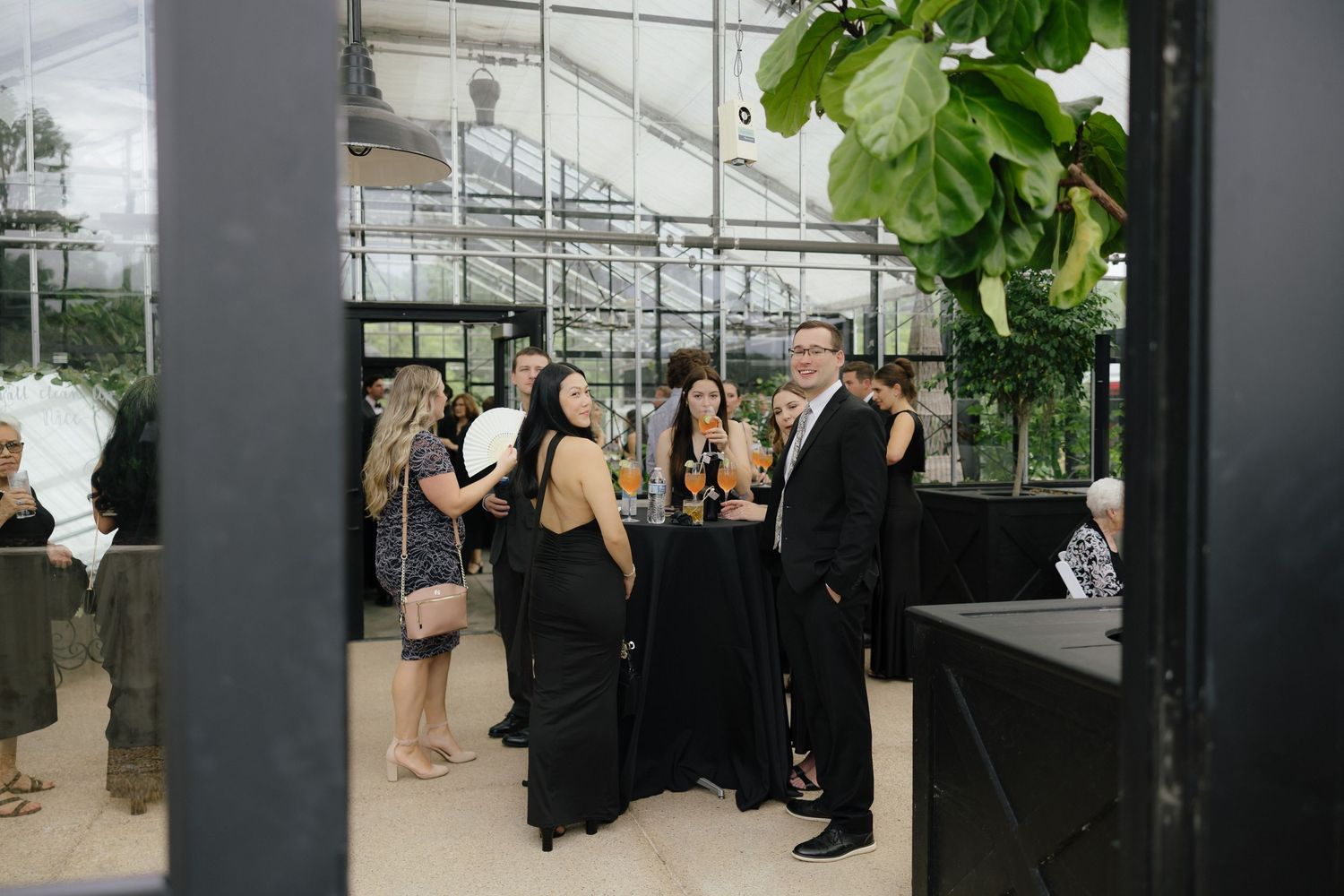 Wedding guests in black formal attire mingling with champagne inside elegant greenhouse cocktail hour.