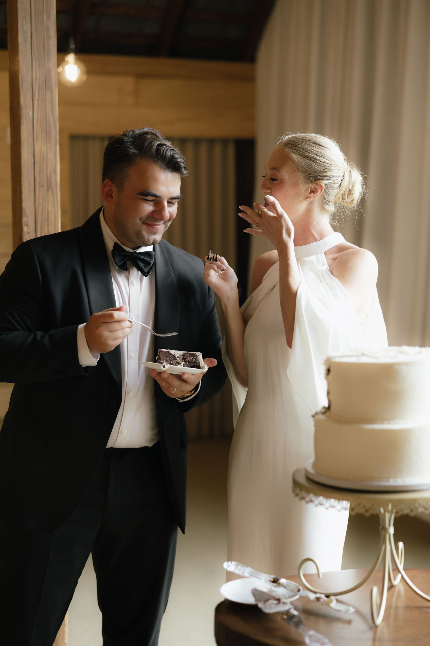 Smiling bride and groom share wedding cake together at elegant rustic reception venue.