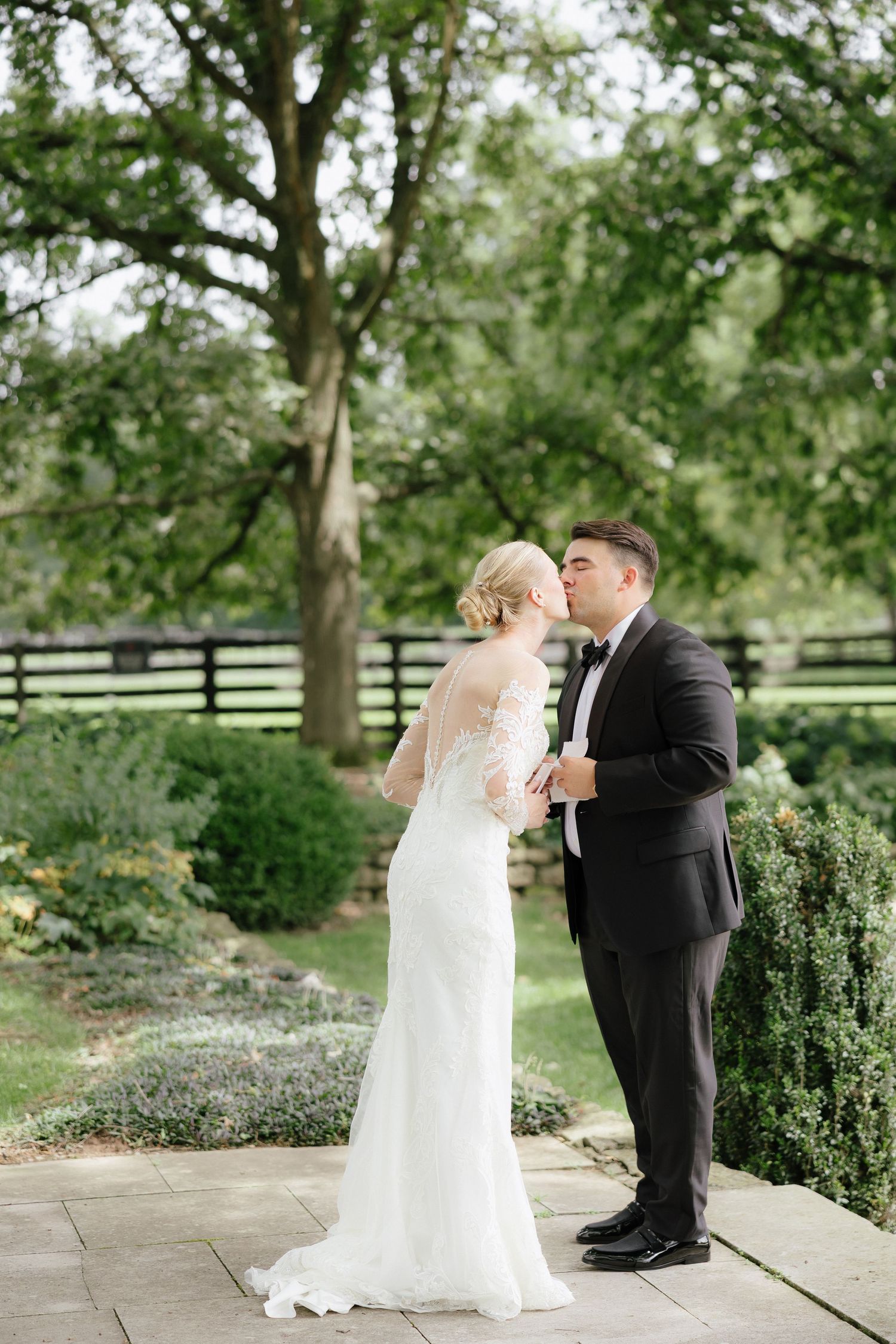 Bride and groom share a romantic kiss outdoors in a lush garden setting, bride wearing elegant lace wedding gown.