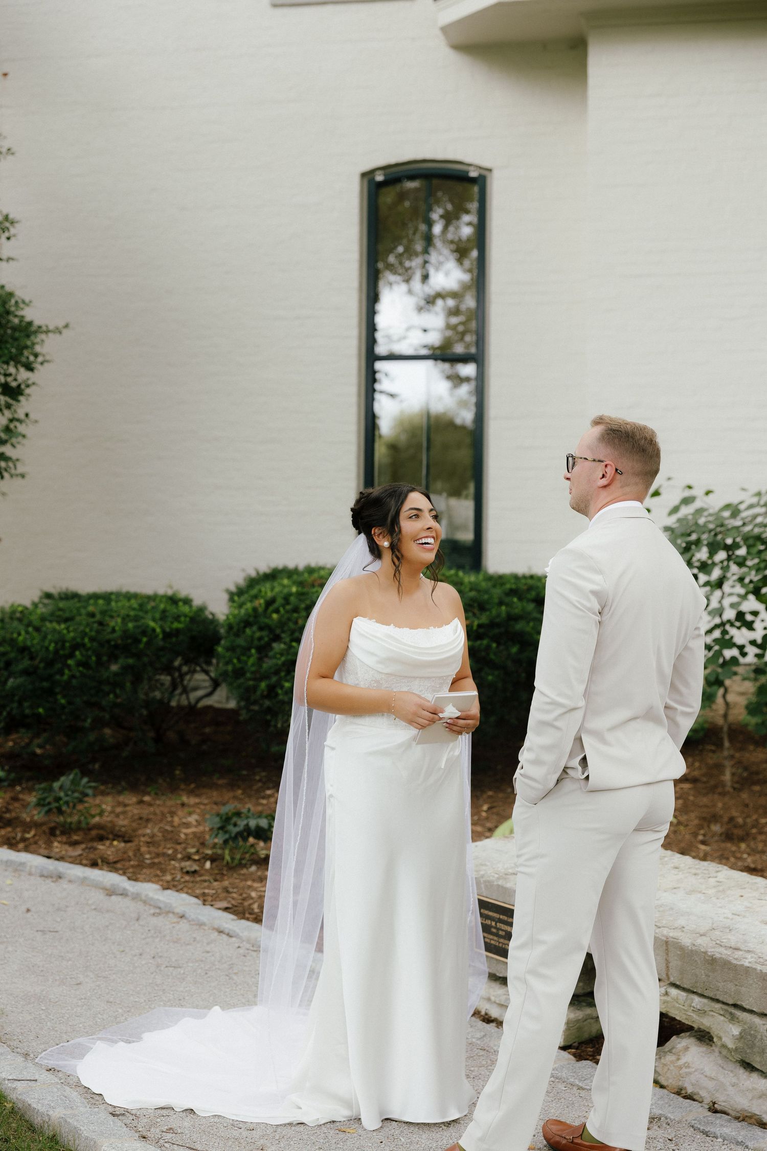 Bride in strapless white gown with veil laughs with groom in ivory suit outside white brick venue.