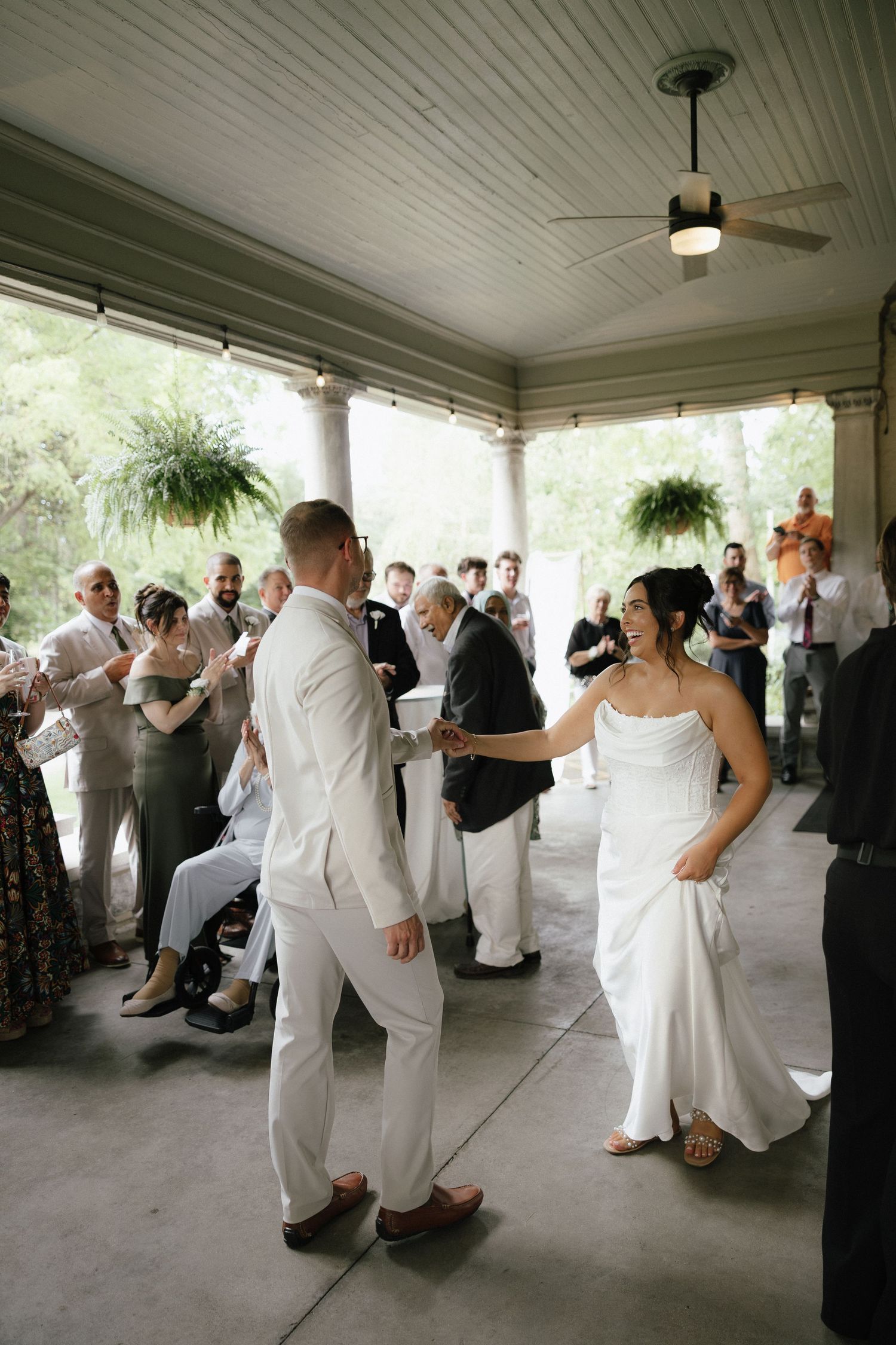 Bride and groom share first dance on covered porch while wedding guests watch and celebrate.