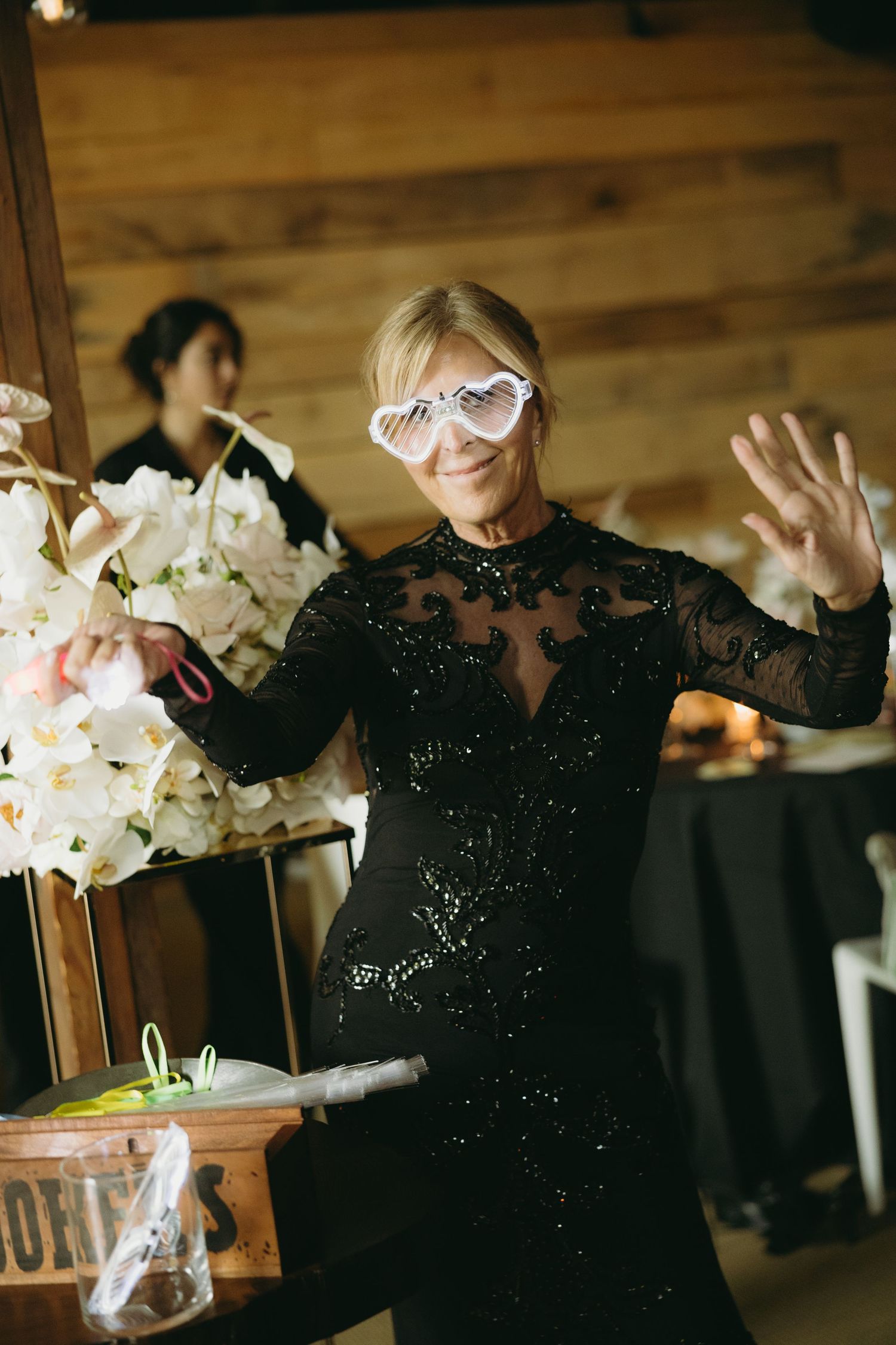 Energetic wedding guest in black sequin dress wearing heart-shaped sunglasses dancing at reception.