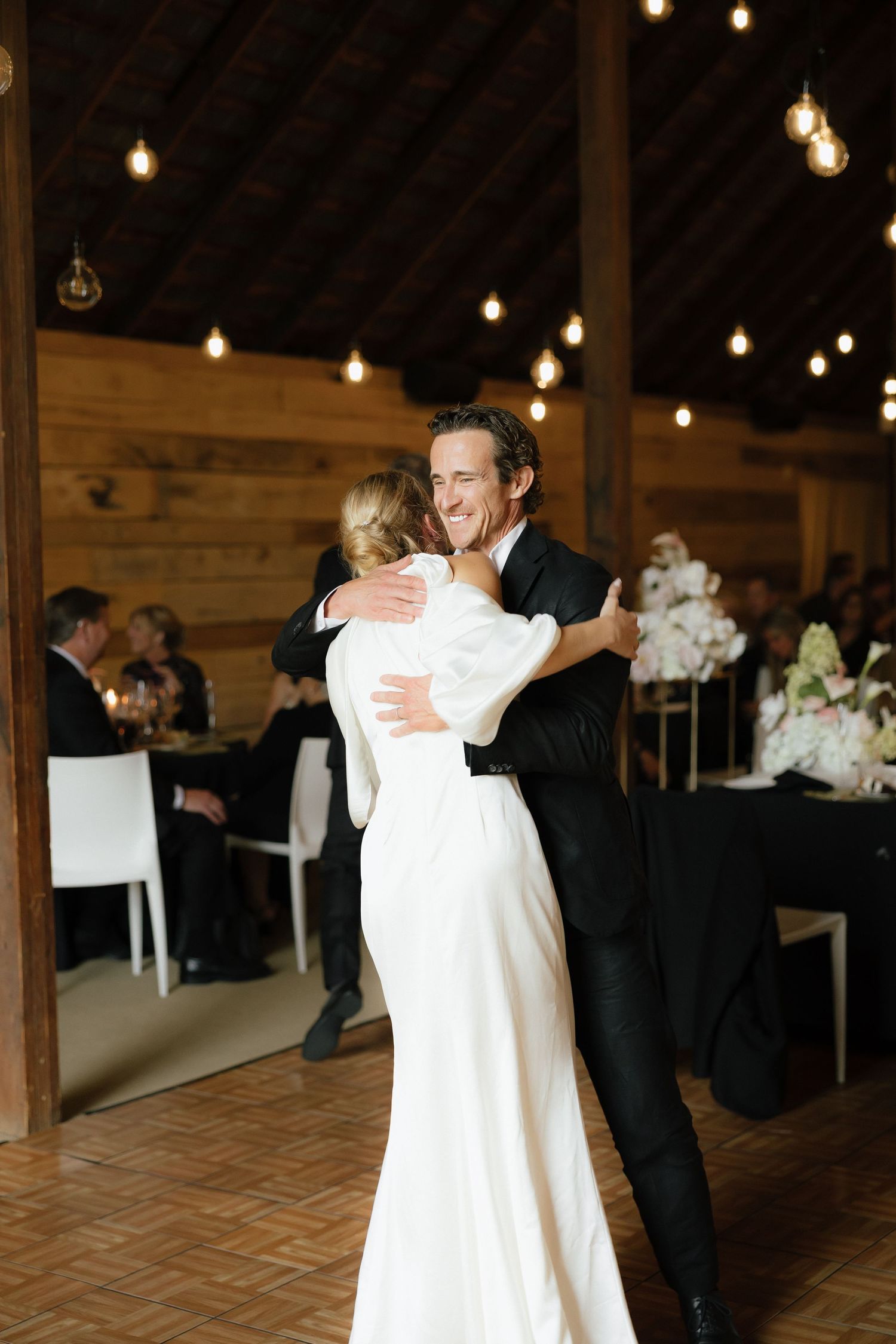 Smiling groom in black tuxedo dances with bride in white gown at rustic barn reception.