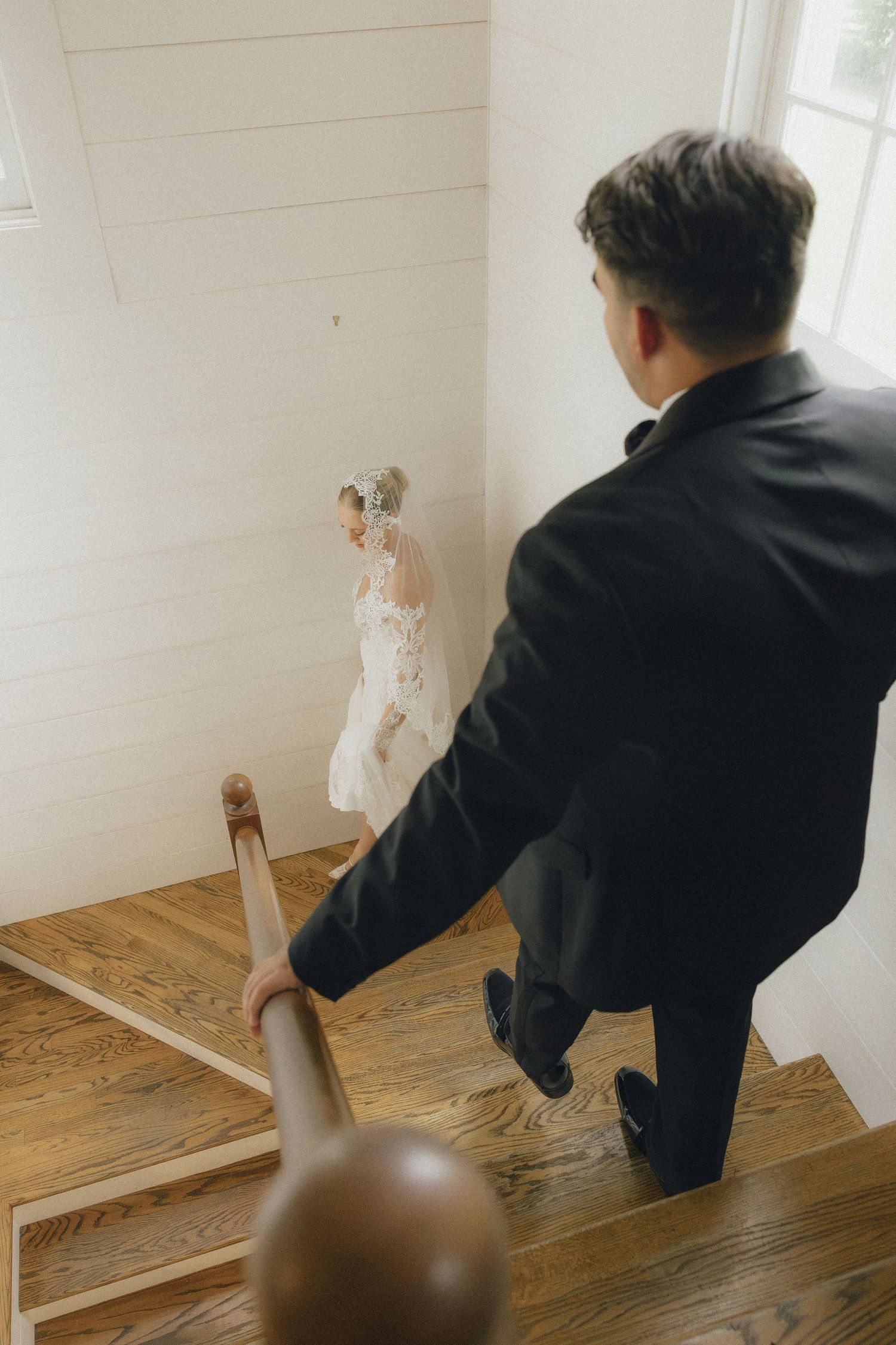 Groom in black suit follows bride in lace gown up wooden staircase inside white wedding venue.