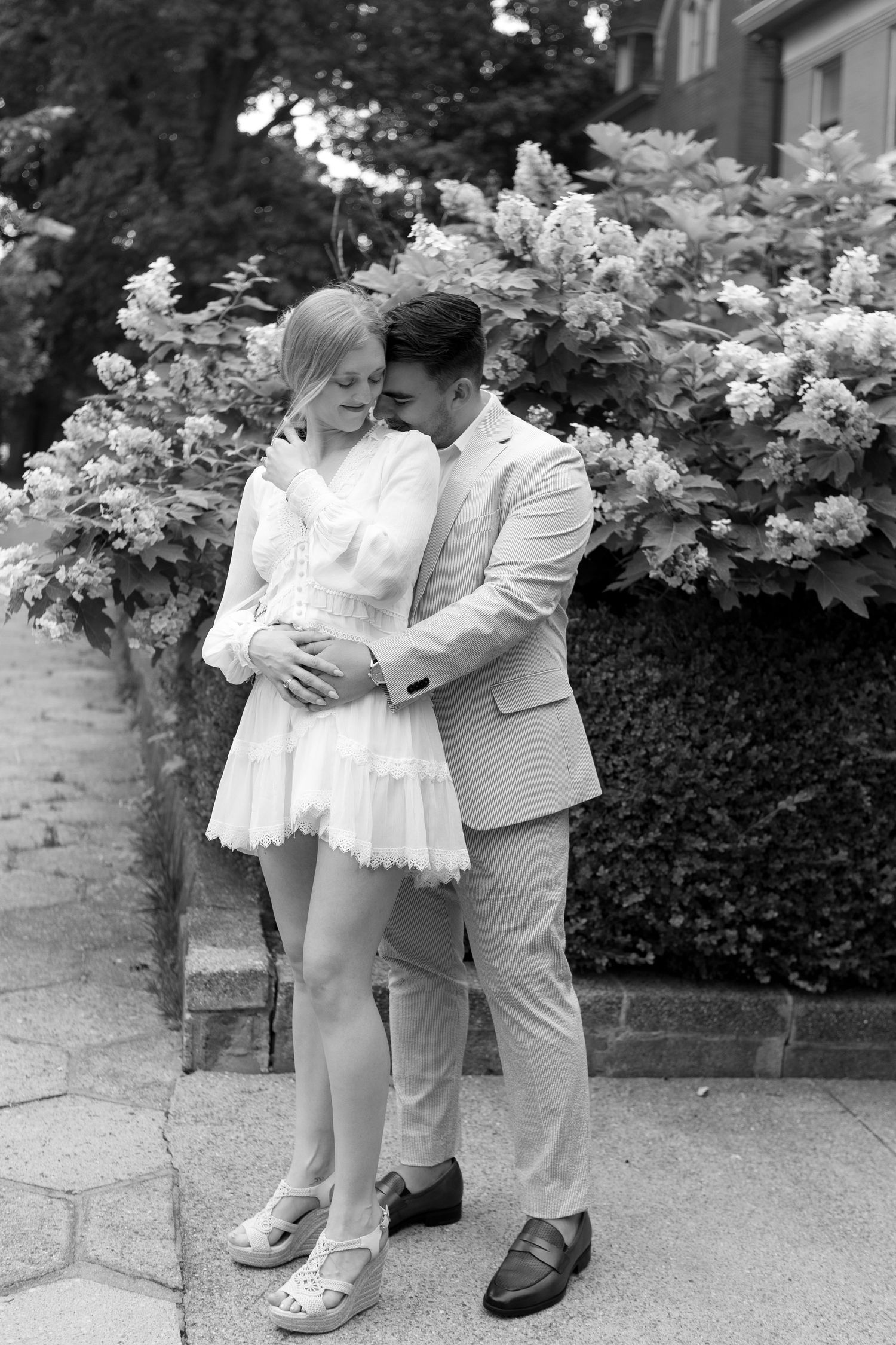 Black and white photo of a couple embracing outdoors near blooming bushes, woman in white dress, man in striped suit.