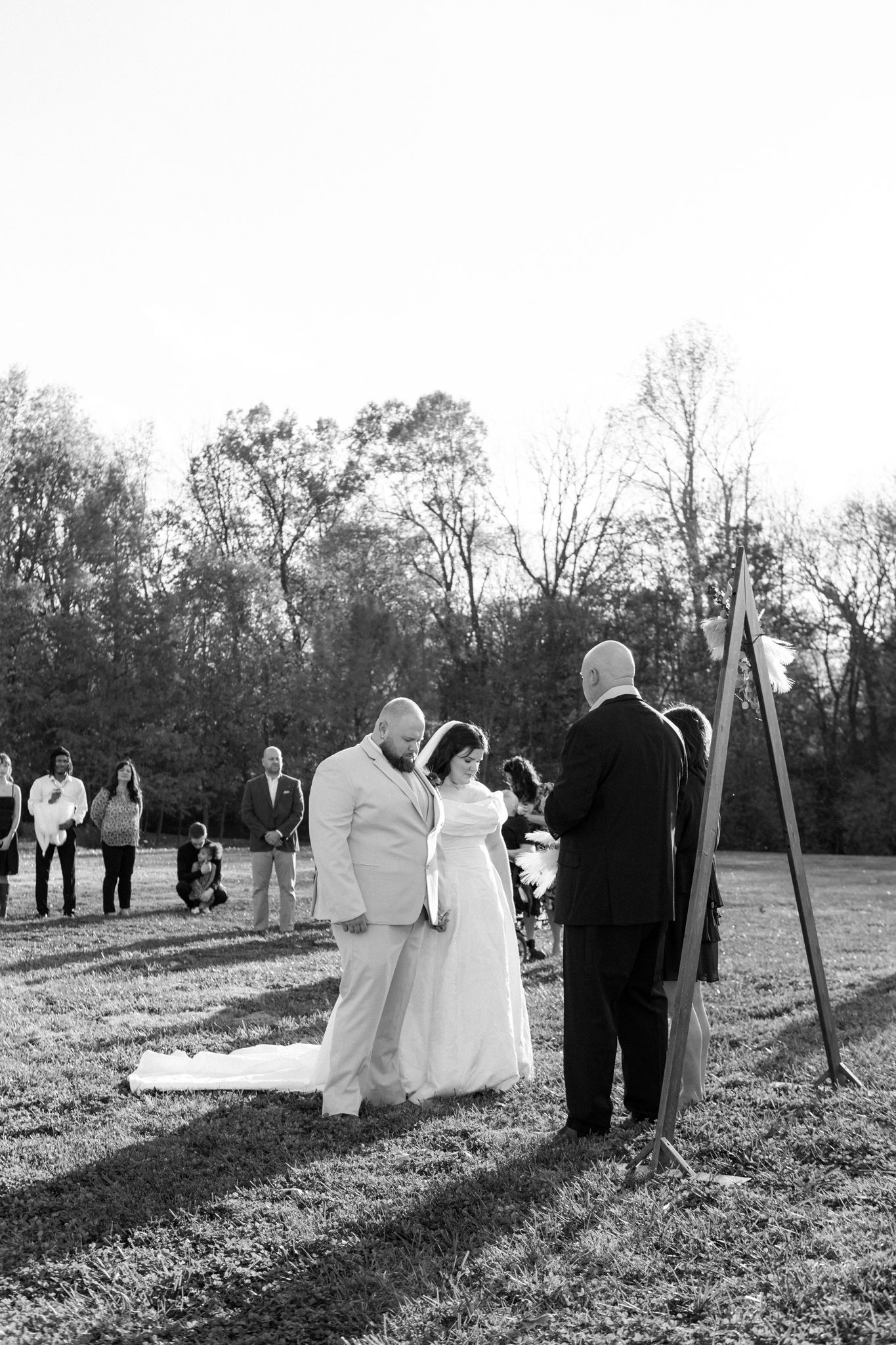 A wedding party walks across a sunlit field in black and white as their shadows stretch across the grass.