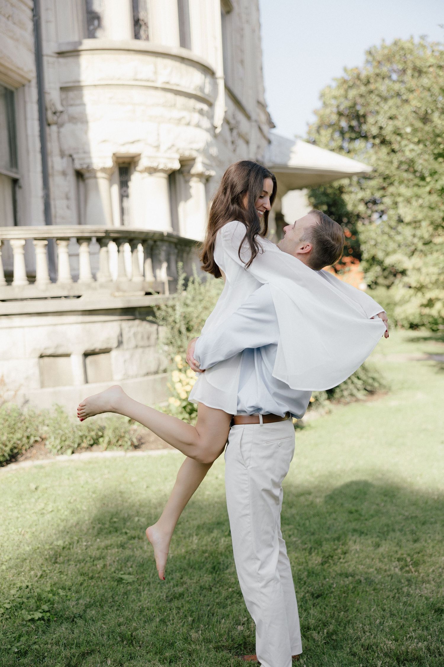 Man lifting woman in white dress outdoors, laughing joyfully in front of a historic stone building.