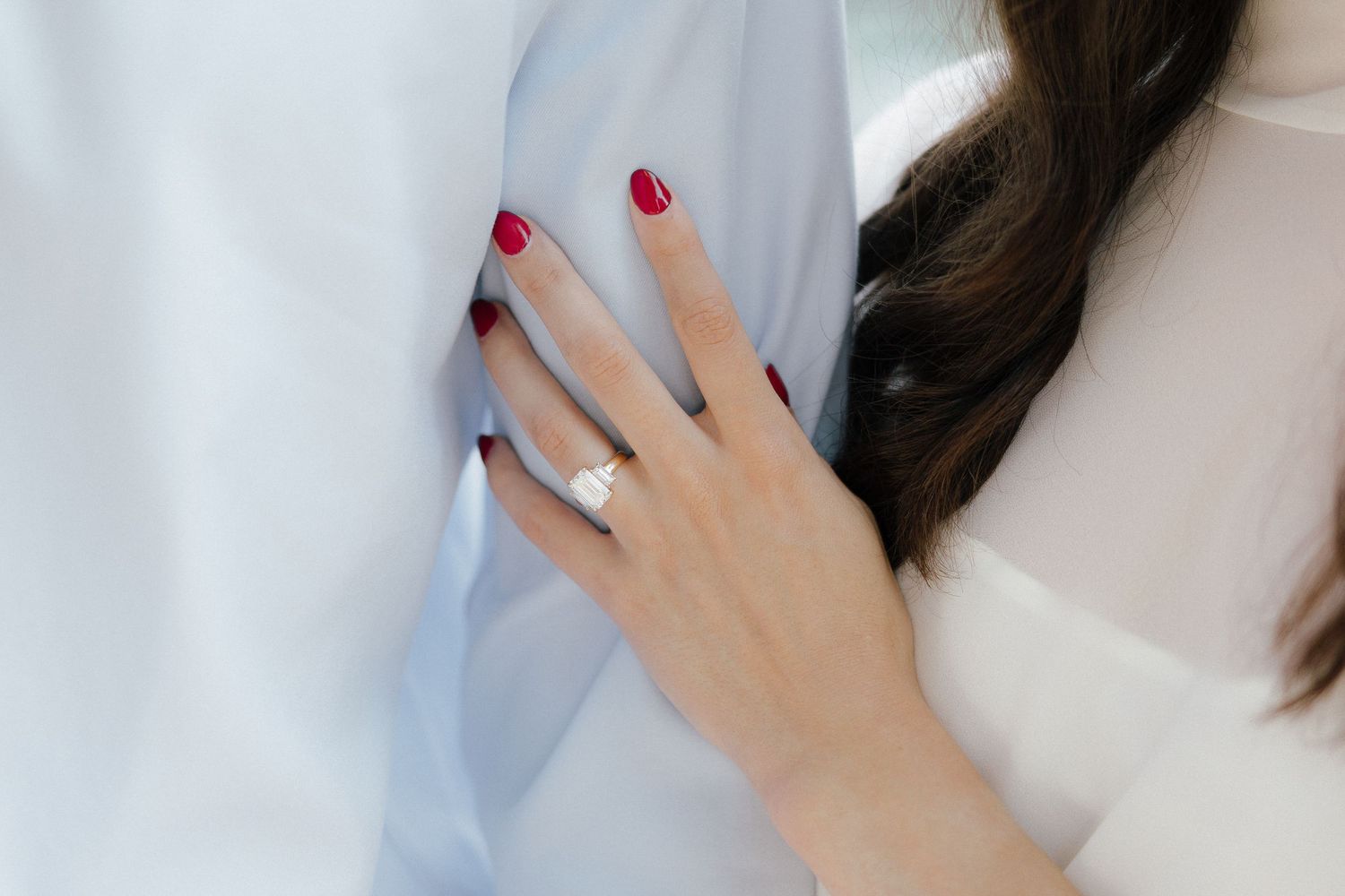 Close-up of engagement ring on woman's hand with red nails resting on man's light blue shirt.