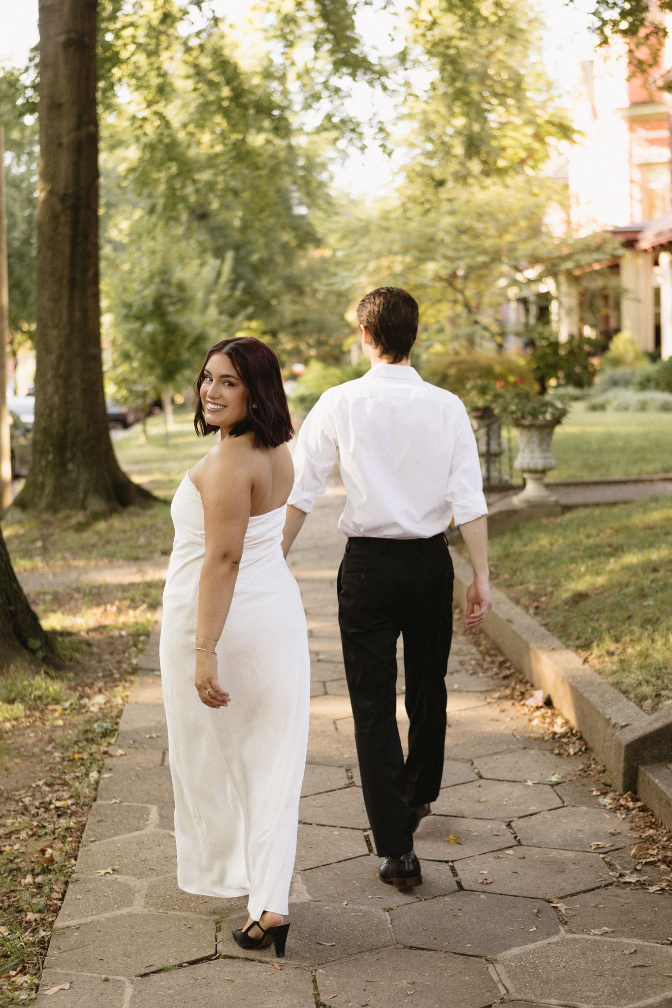 A couple in formal attire walks along a stone path through a garden setting during golden hour.