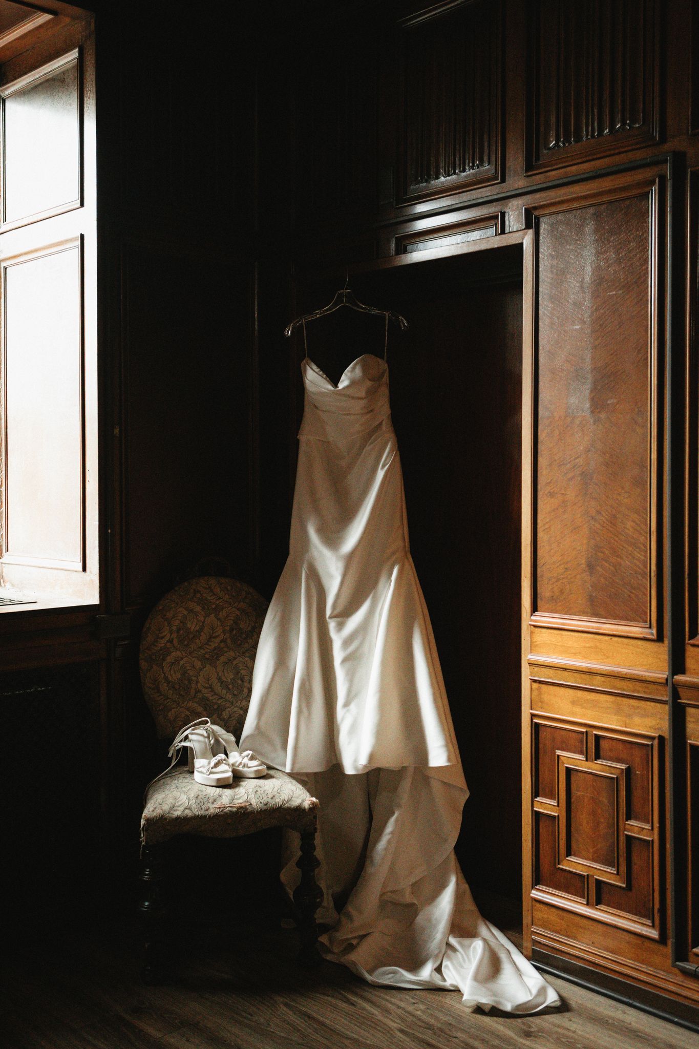 Wedding dress hanging in a dark wooden room with dramatic natural lighting through windows.
