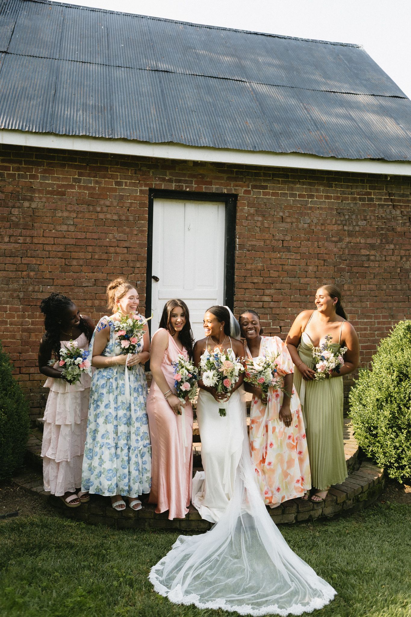 A bridal party in pastel dresses poses together against a rustic brick barn on a grassy lawn.