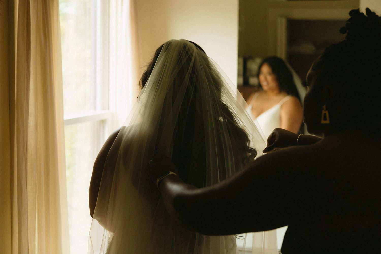 A bride in a white veil stands near a window during wedding preparations as soft natural light filters through.