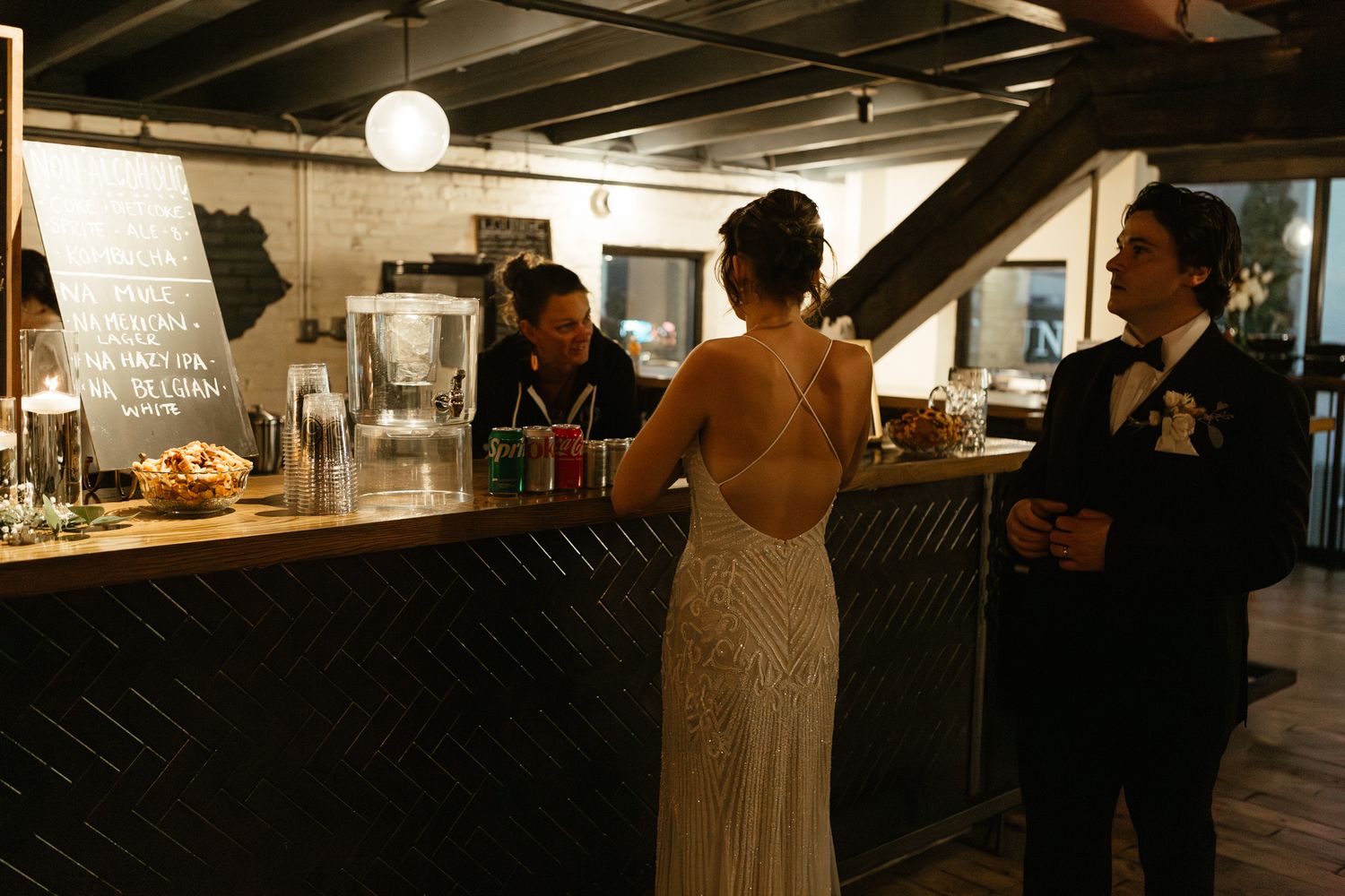 Wedding guests mingle at a dimly lit bar area during an evening reception.