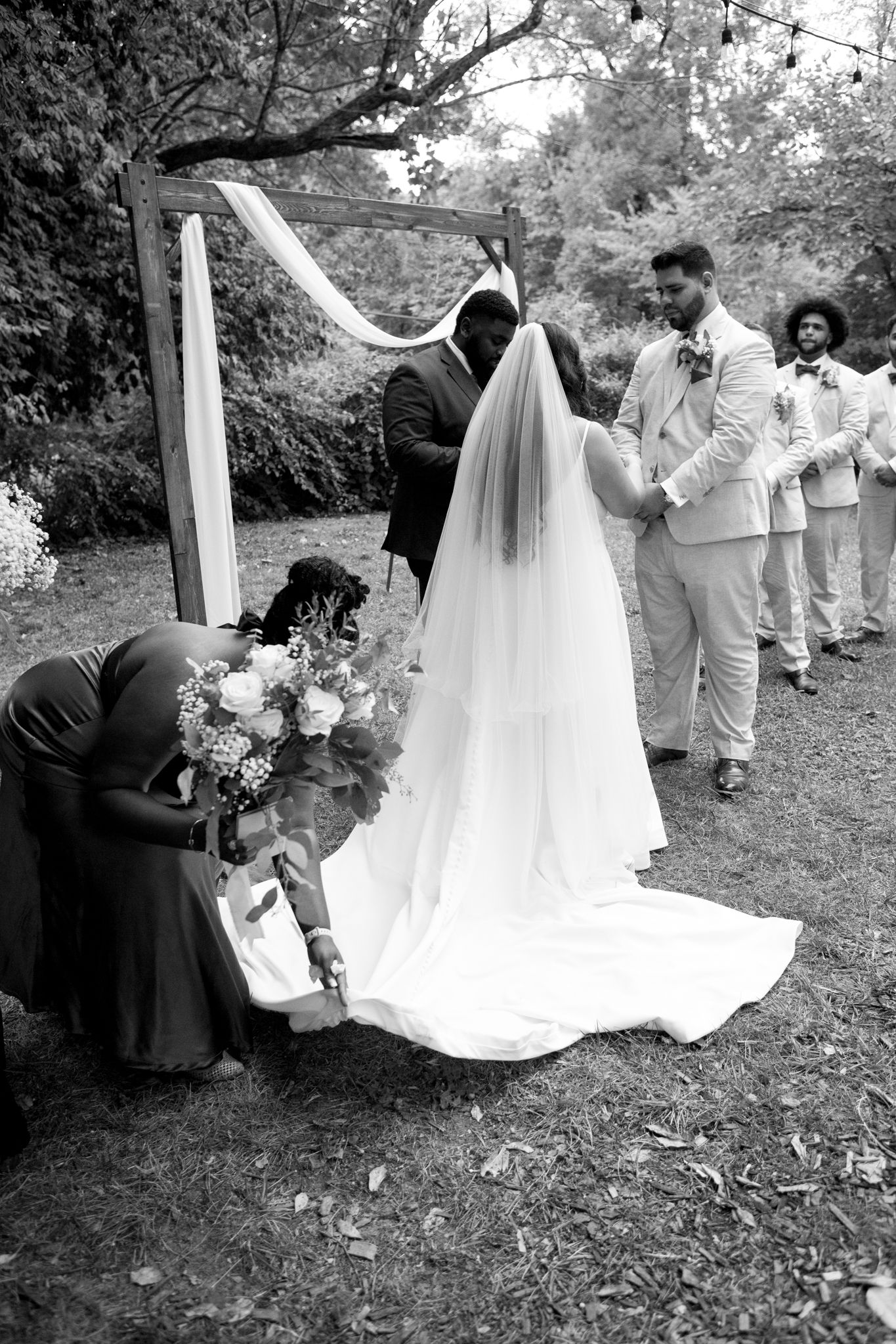 A black and white outdoor wedding ceremony under an arch with attendants and a flowing long white veil in a garden setting.