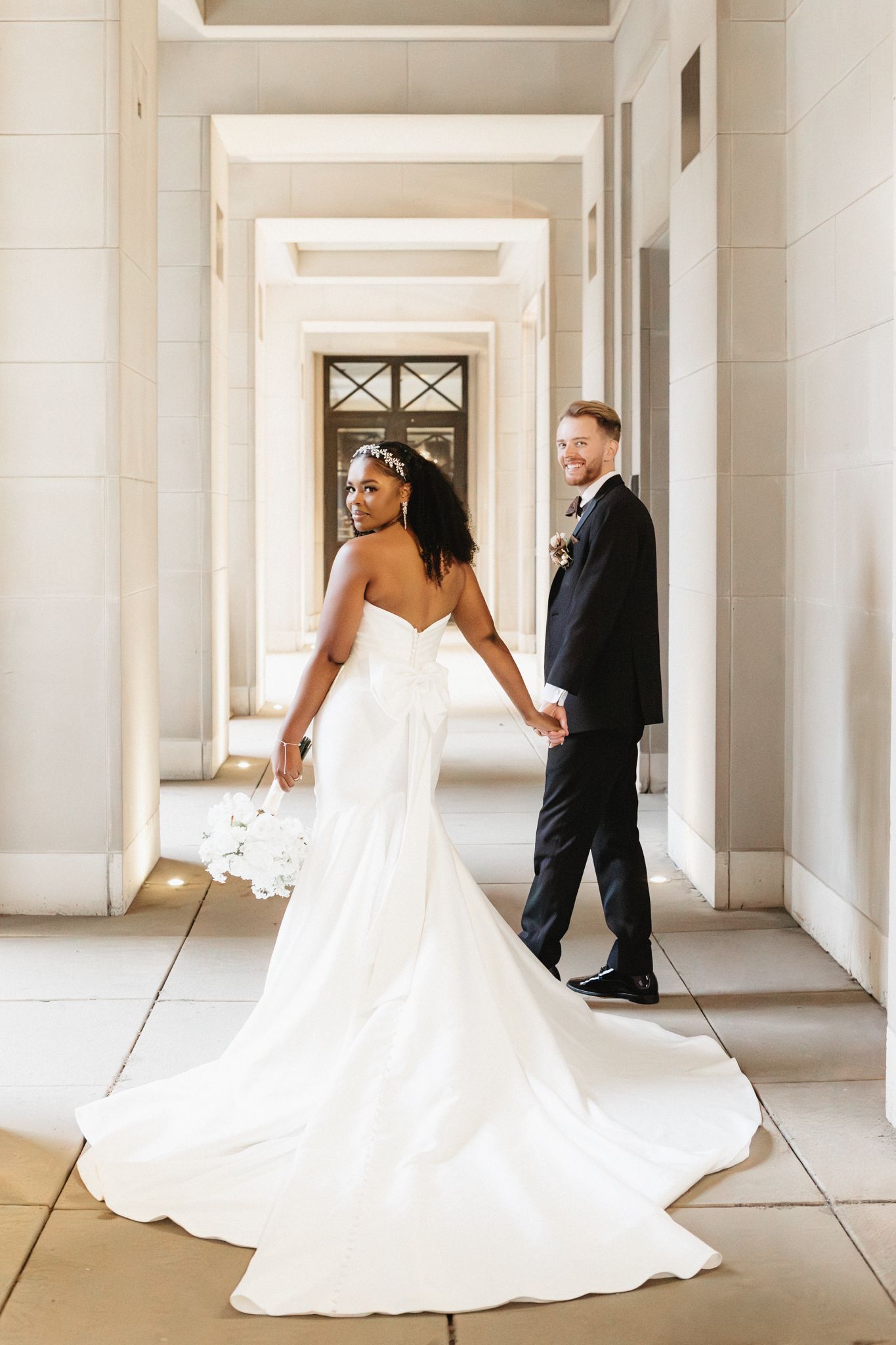 Bride's flowing white dress trails behind as couple walks through columned hallway.