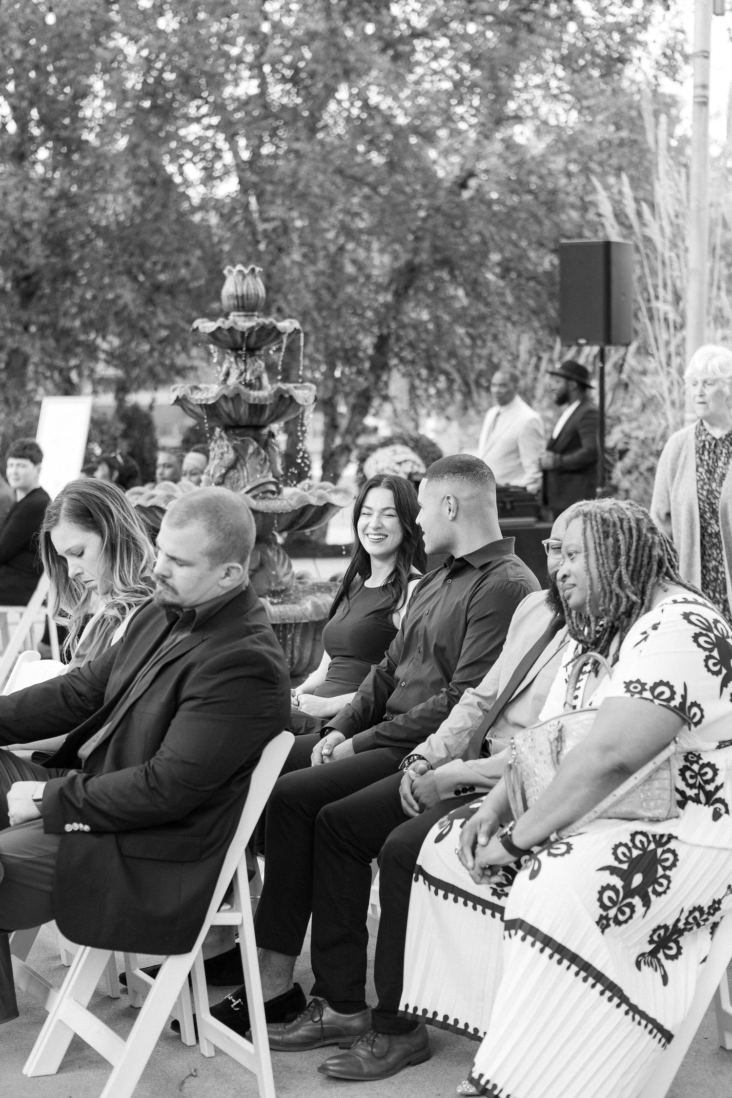 Wedding ceremony guests seated on white chairs in an outdoor garden setting.