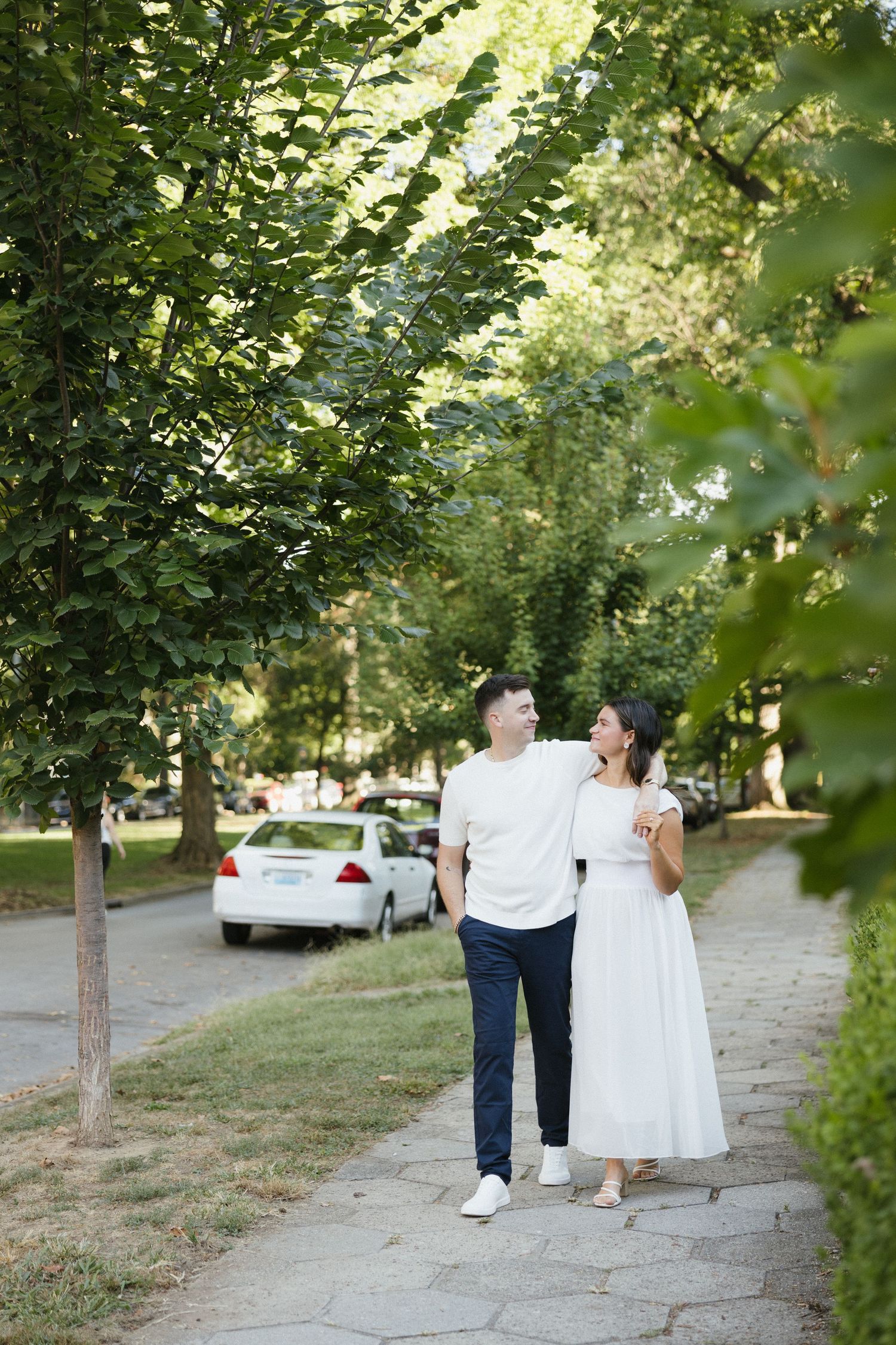 Couple walking hand in hand along a tree-lined park path, man in white shirt and woman in white dress.