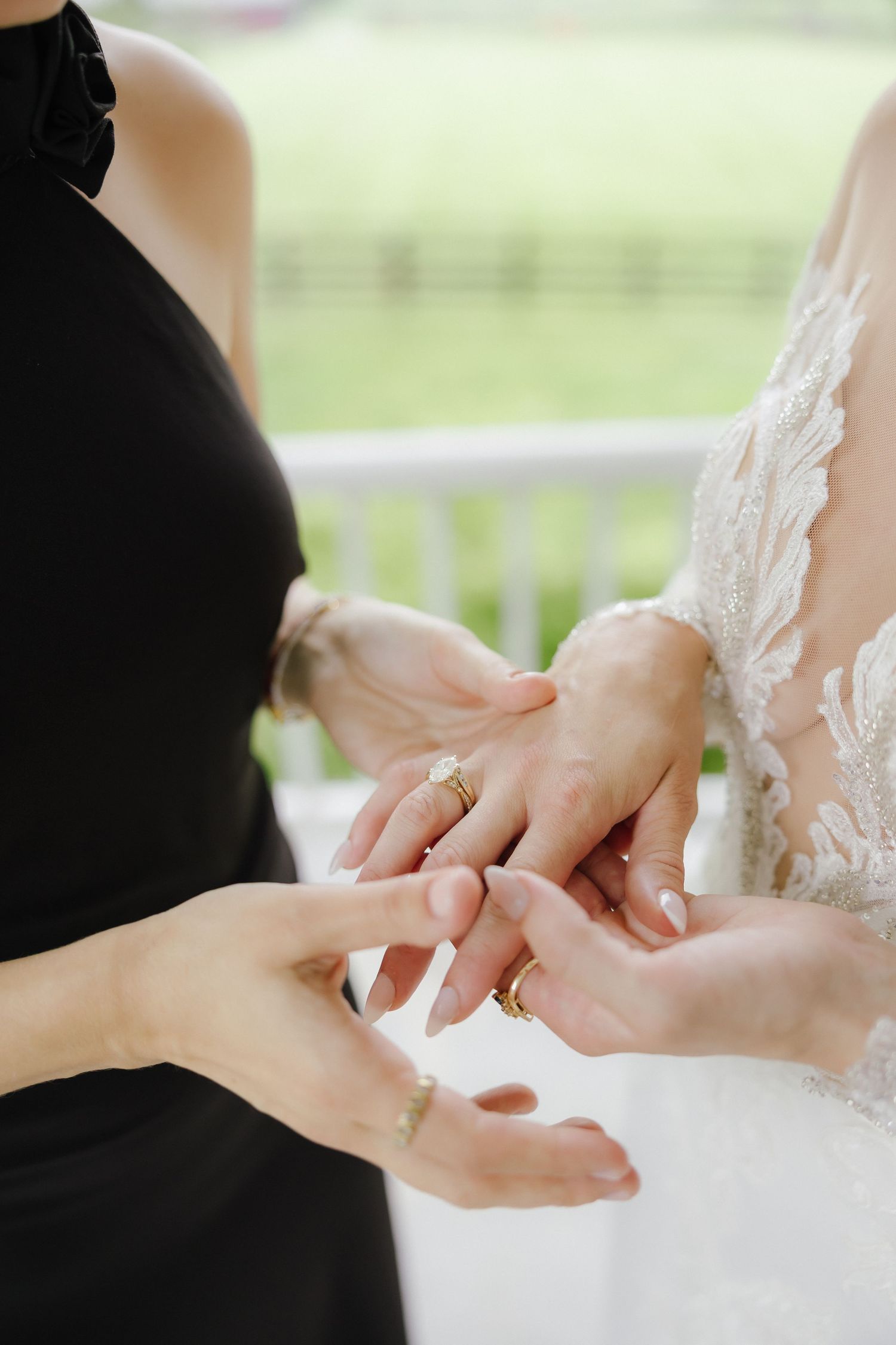 Bridesmaid in black dress helps bride in lace gown with wedding ring on outdoor porch.
