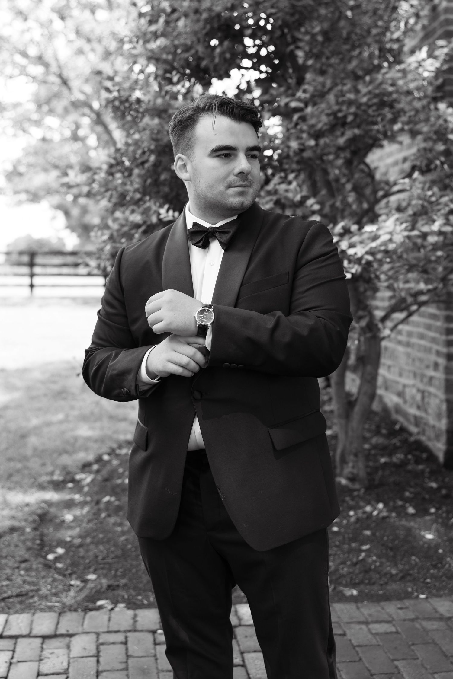 Black and white photo of a well-dressed man in a tuxedo adjusting his cufflinks outdoors near trees.