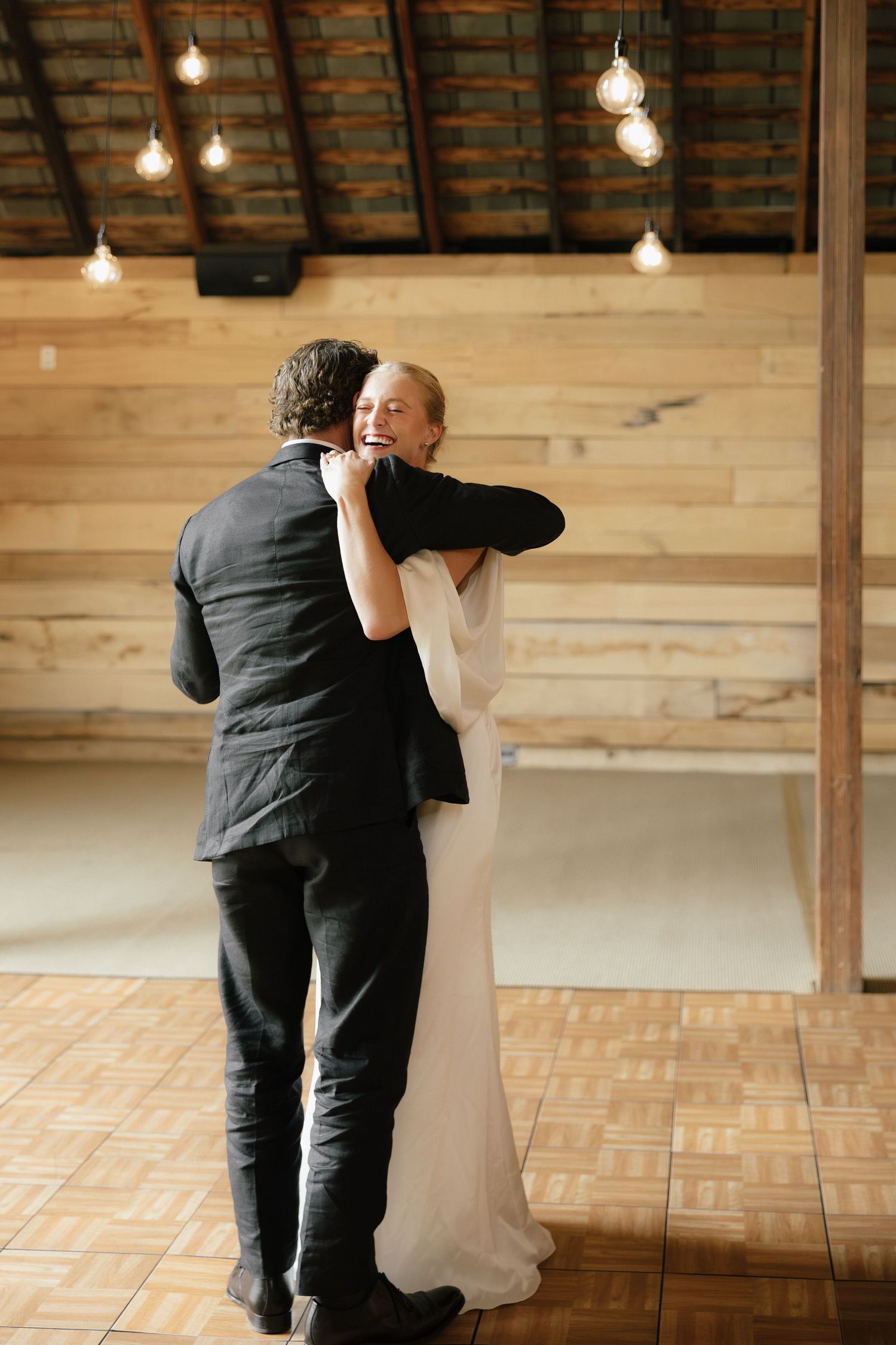 Bride and groom sharing a first dance embrace on a wooden dance floor in a warmly lit rustic barn venue.