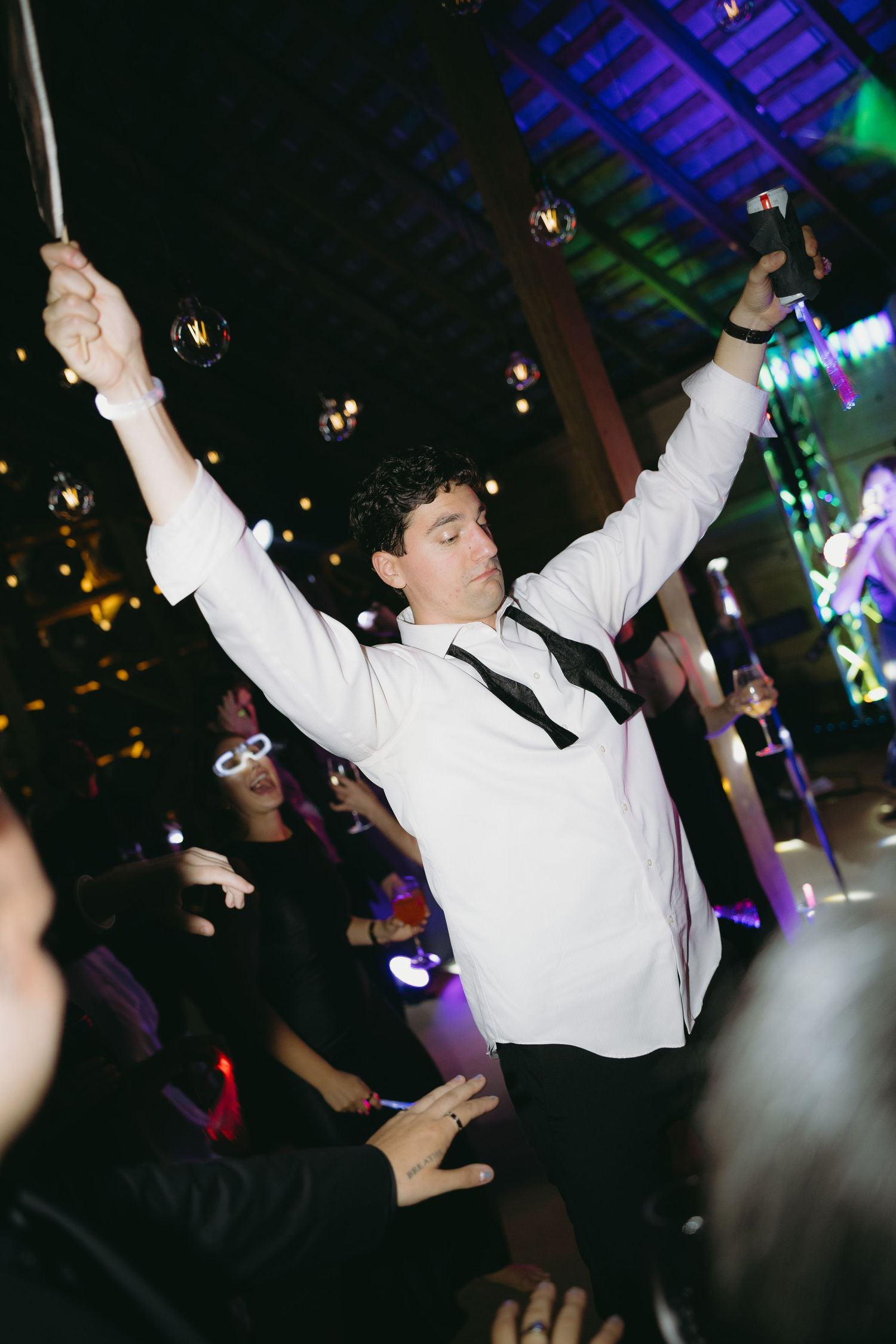Groom celebrates on dance floor with arms raised at lively wedding reception with colorful lights.