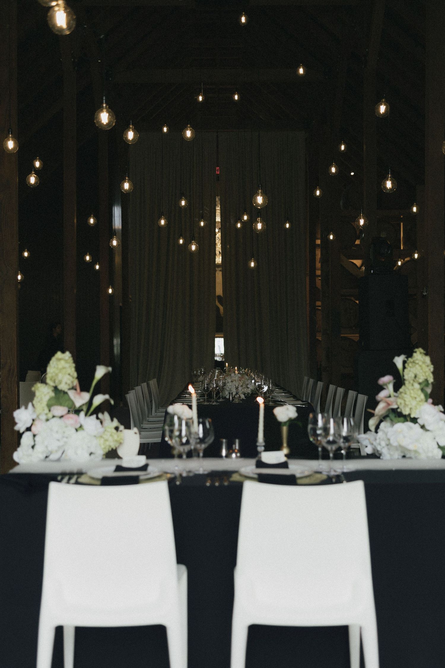 Elegant wedding reception table with black linens, white chairs, floral centerpieces, and Edison bulb lighting.