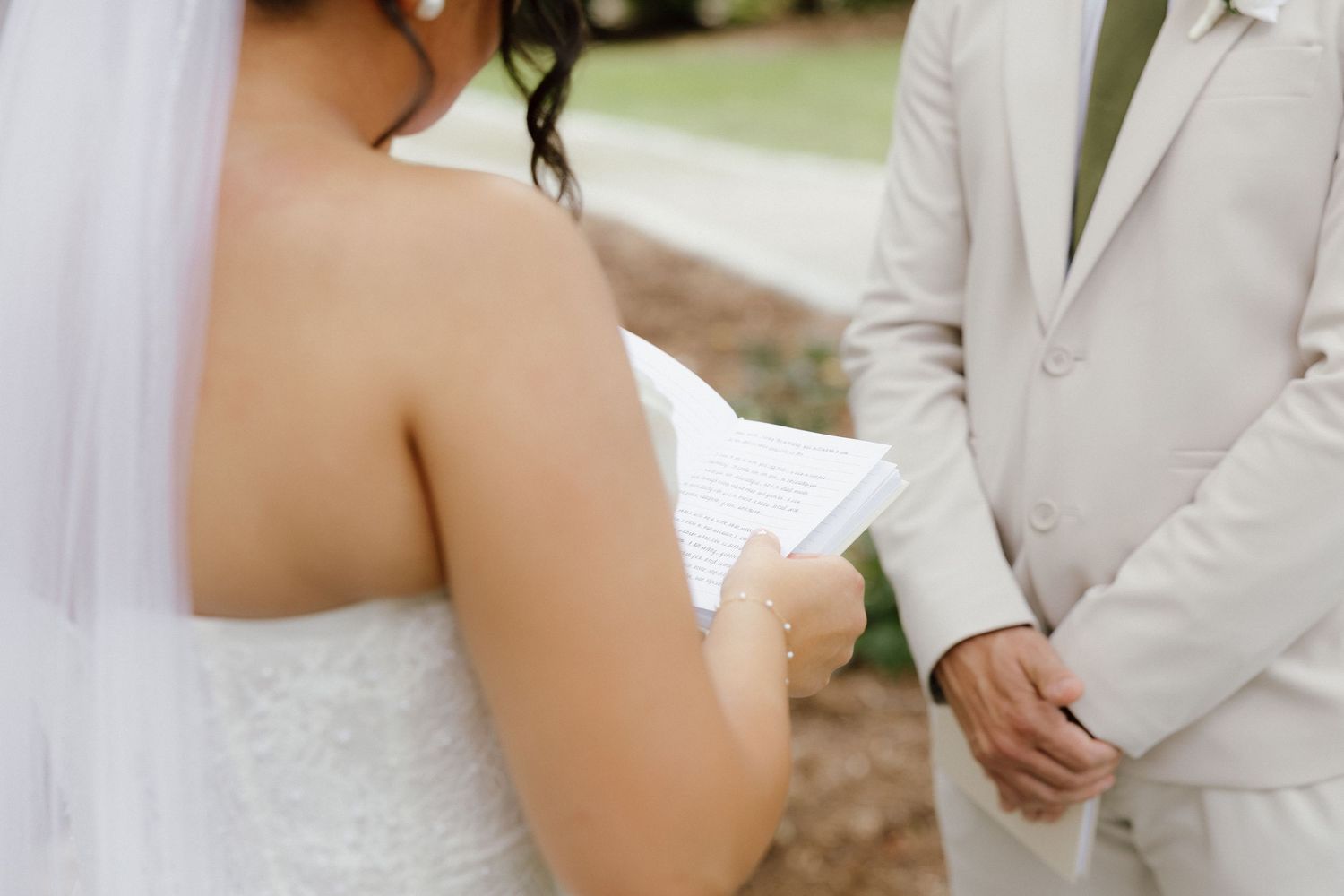 Bride reading handwritten vows to groom in beige suit during outdoor wedding ceremony.