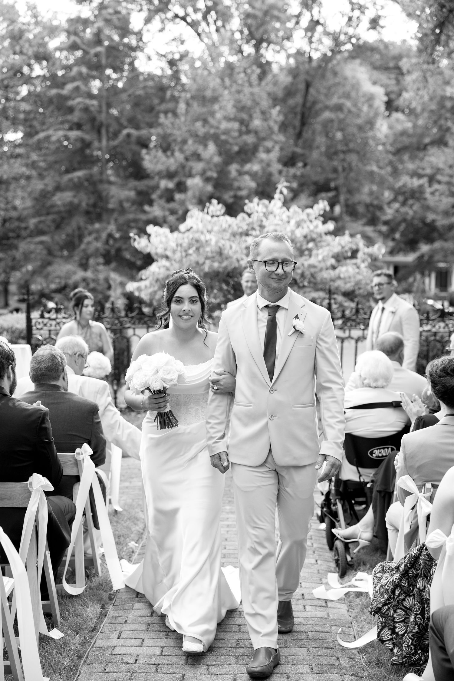 Bride and groom walk back down aisle after wedding ceremony surrounded by seated guests outdoors.