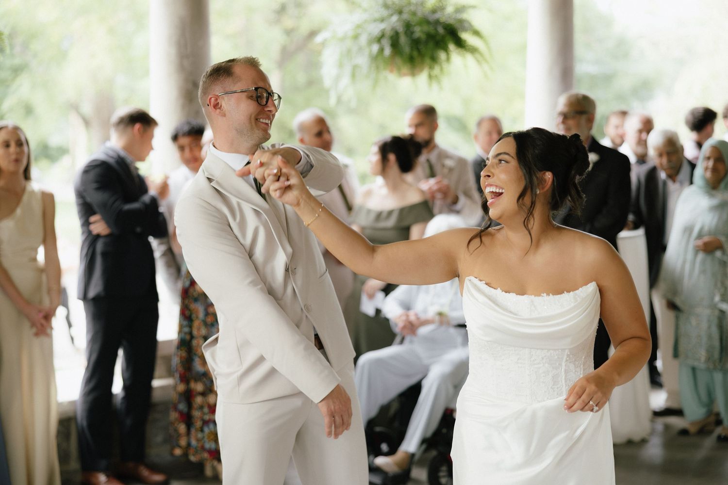 Bride and groom share joyful first dance under covered outdoor pavilion surrounded by wedding guests.