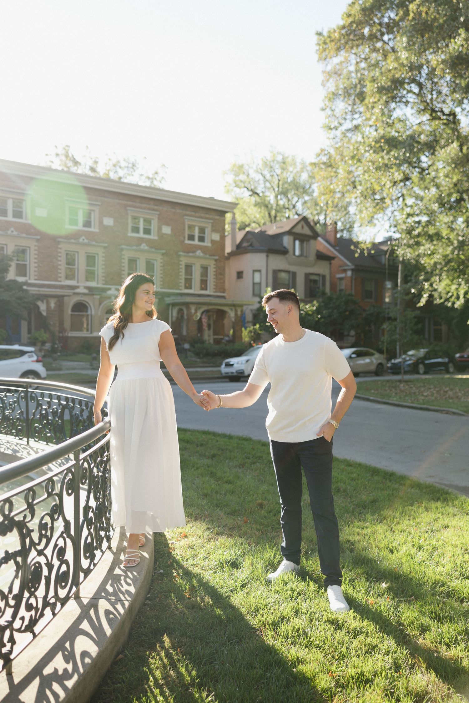 Couple holding hands on ornate bridge in sunny park with historic brick homes in background.